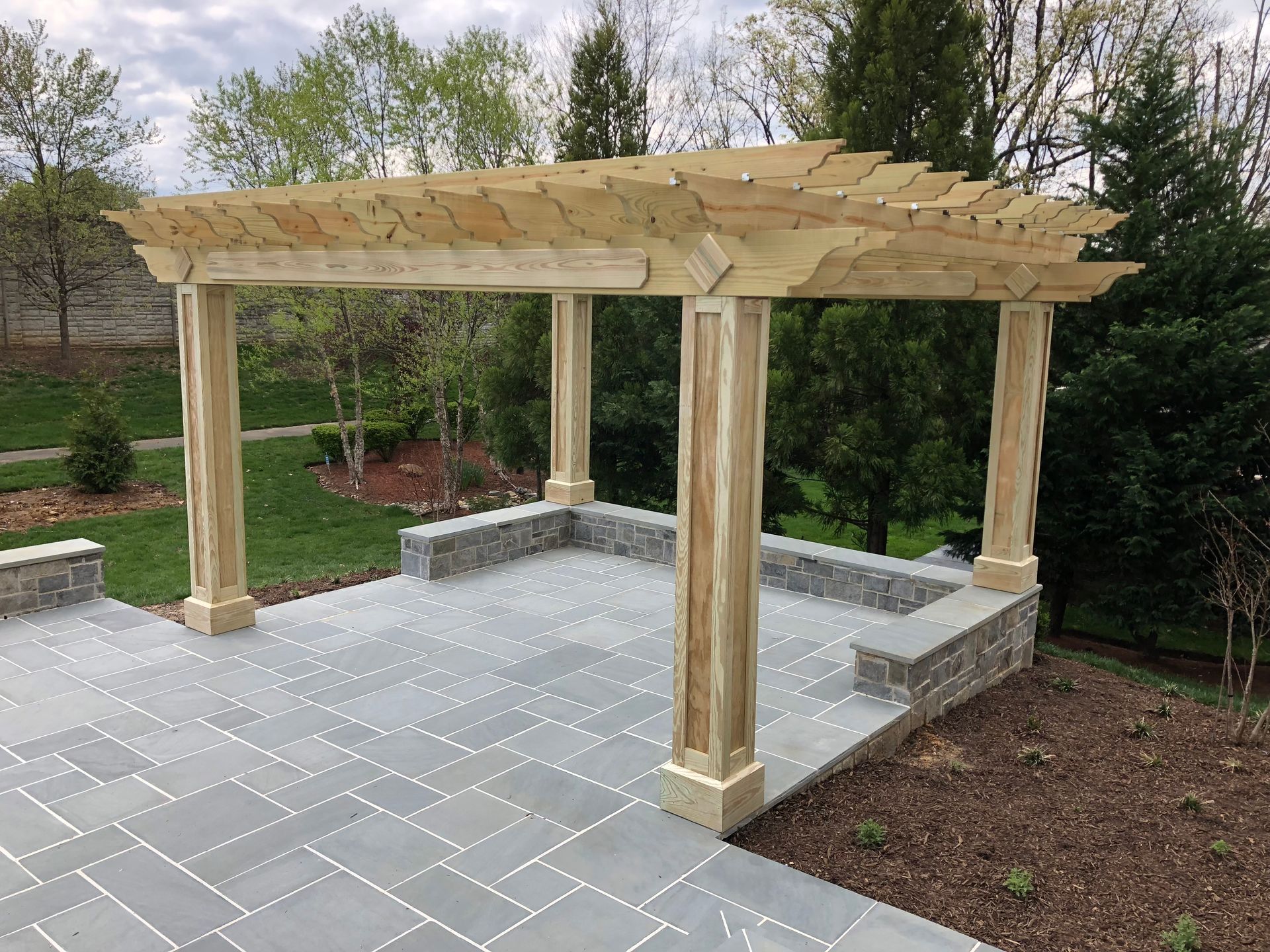 Pergola with wooden beams, supported by columns, on a stone patio with a low wall; trees in background.