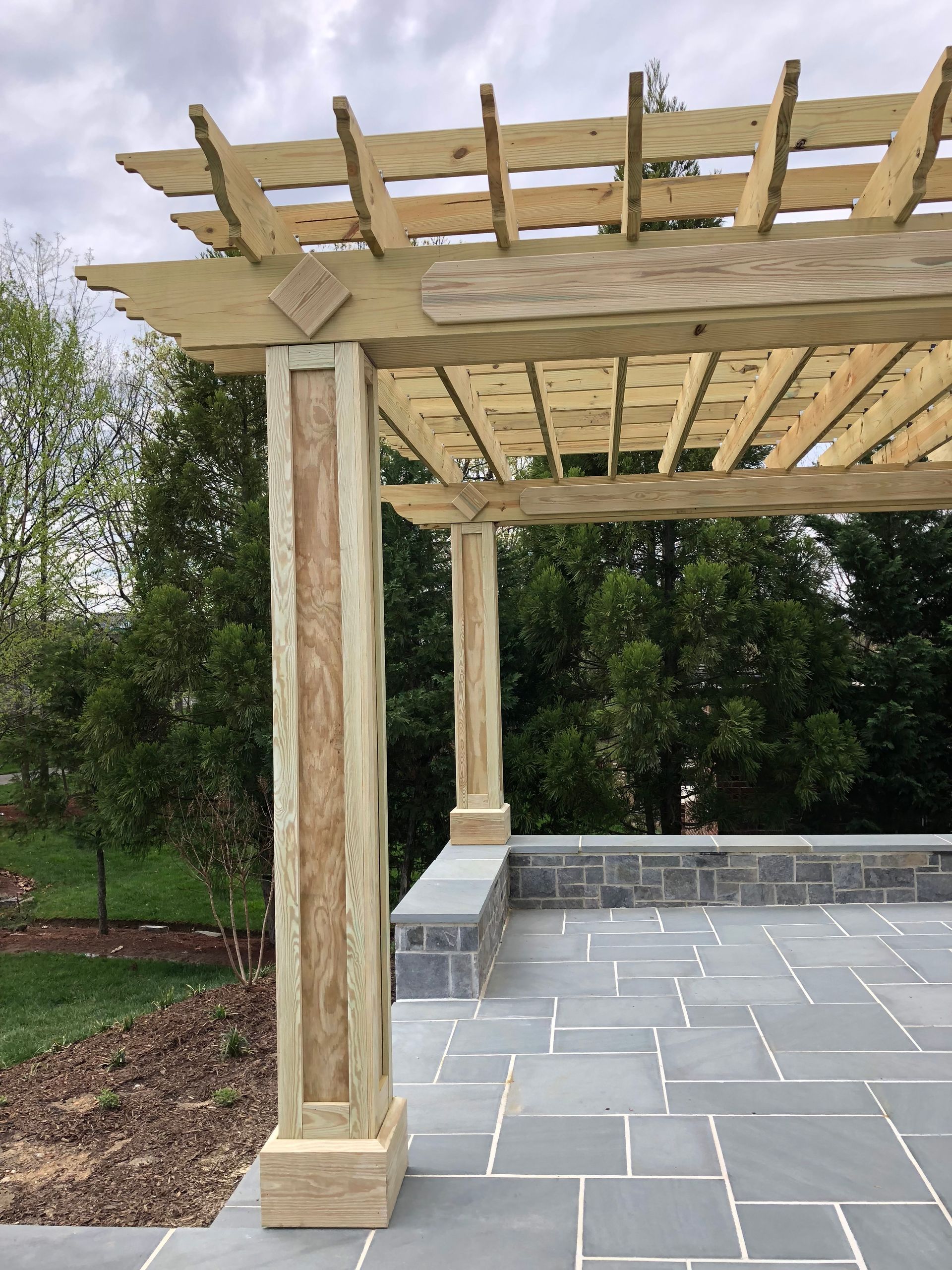 Wooden pergola with decorative columns on a stone patio overlooking greenery.