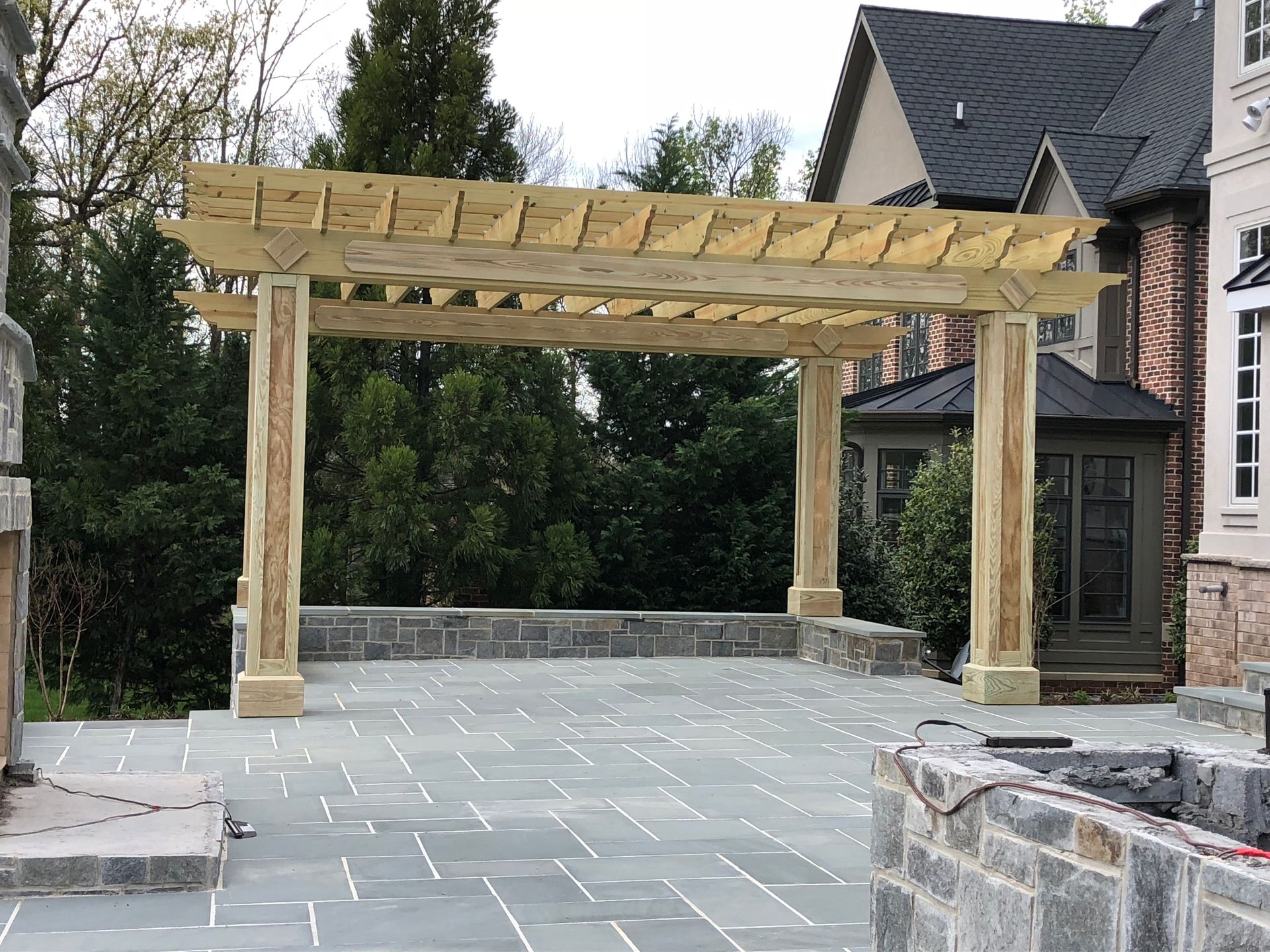 Pergola over a stone patio with a house in the background. Wooden posts and beams.