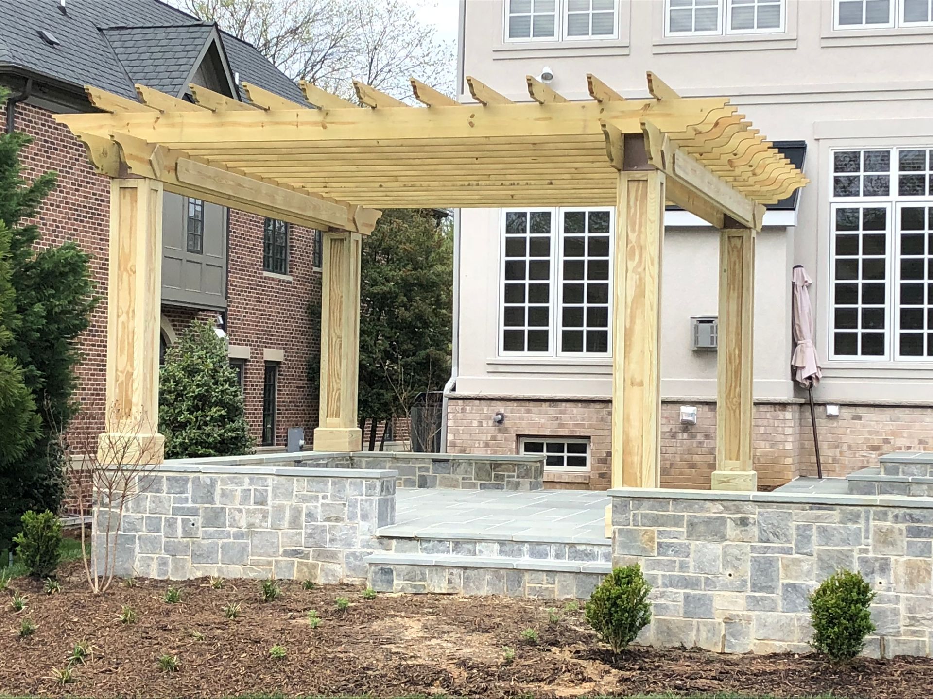 Wooden pergola over a stone patio with steps, next to a beige house with large windows.