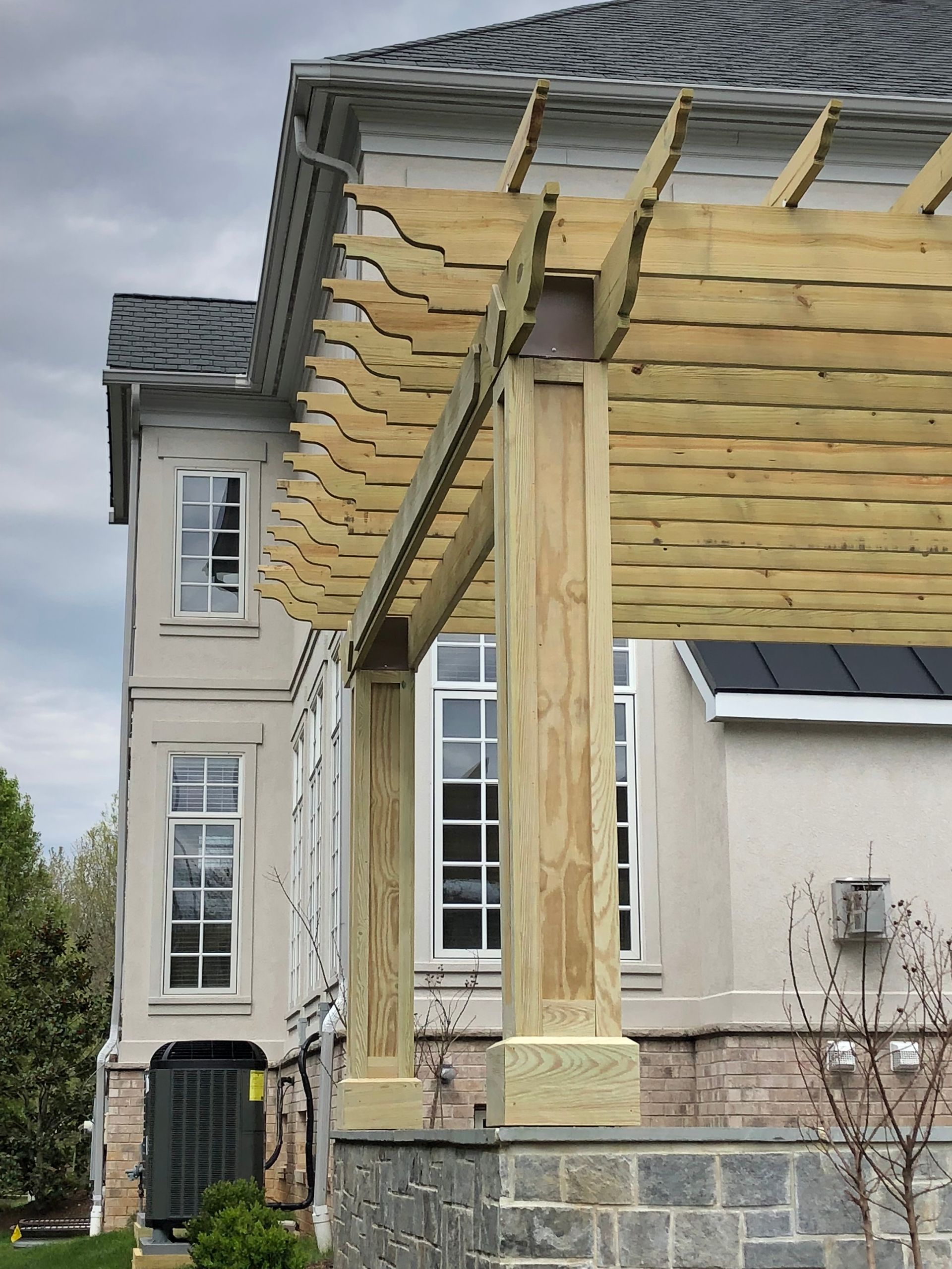 Pergola with wooden beams attached to a light-colored house with a dark roof and multiple windows.