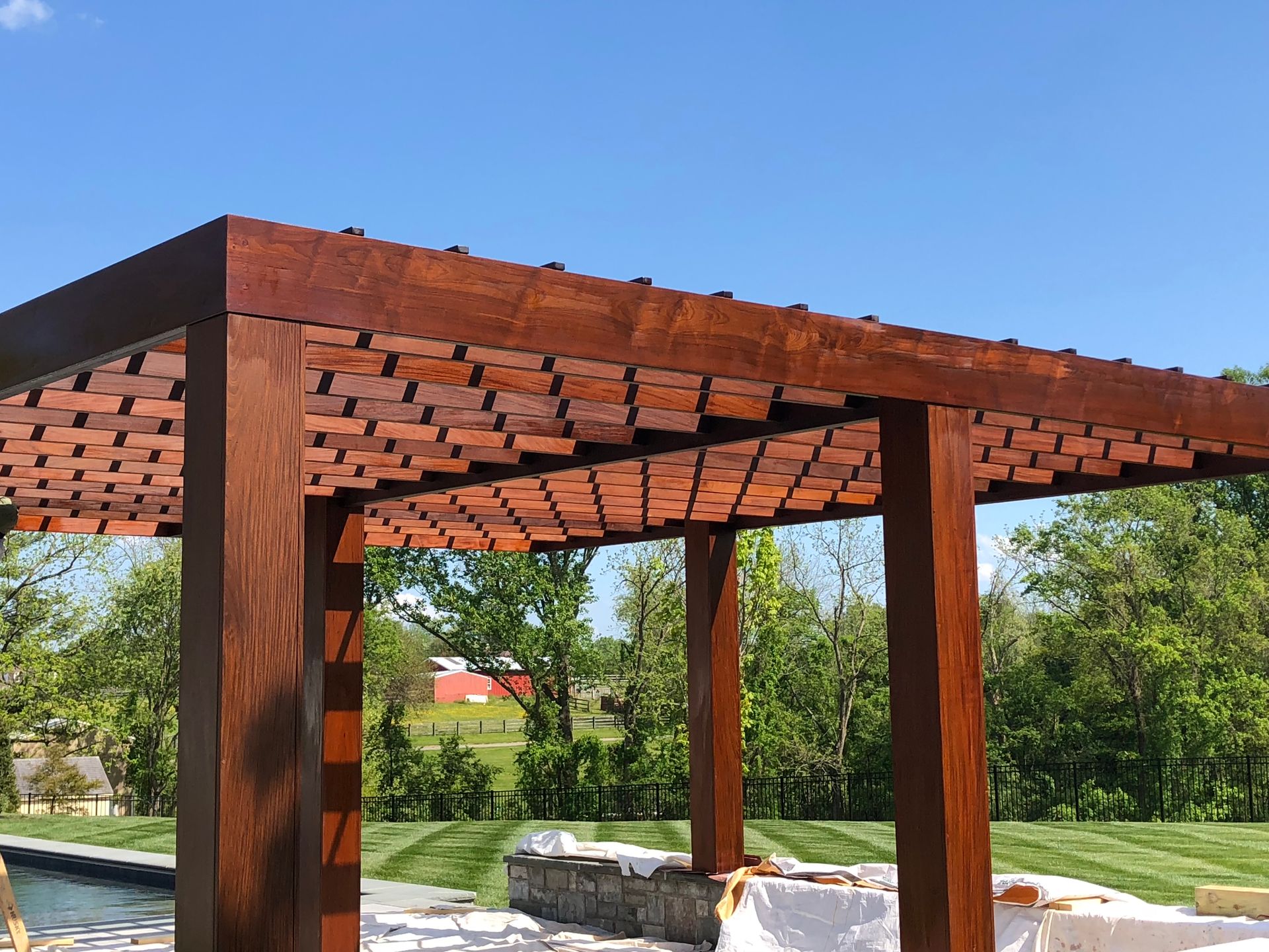 Wooden pergola with brick-like slats, set against a blue sky, near a pool and grassy yard.