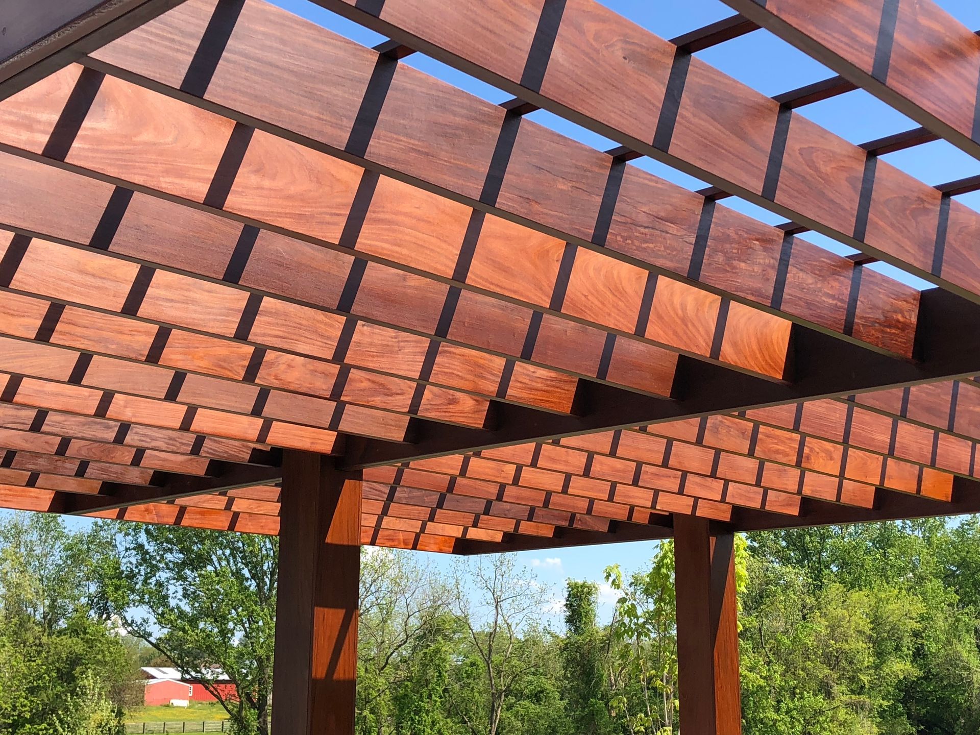 Wooden pergola roof with square blocks casting shadows, set against a blue sky and trees.