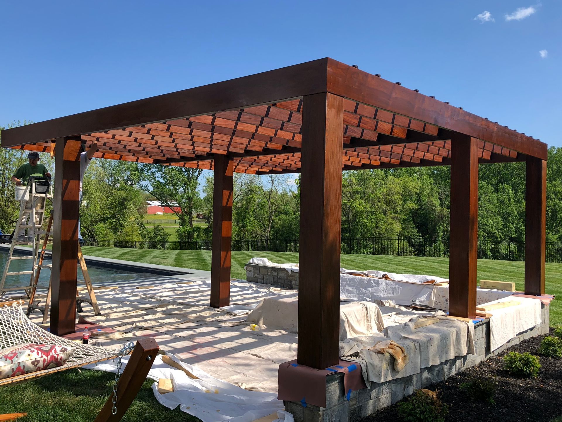 Wooden pergola with brick-patterned roof, built near a pool; a person paints the structure in a sunny outdoor setting.