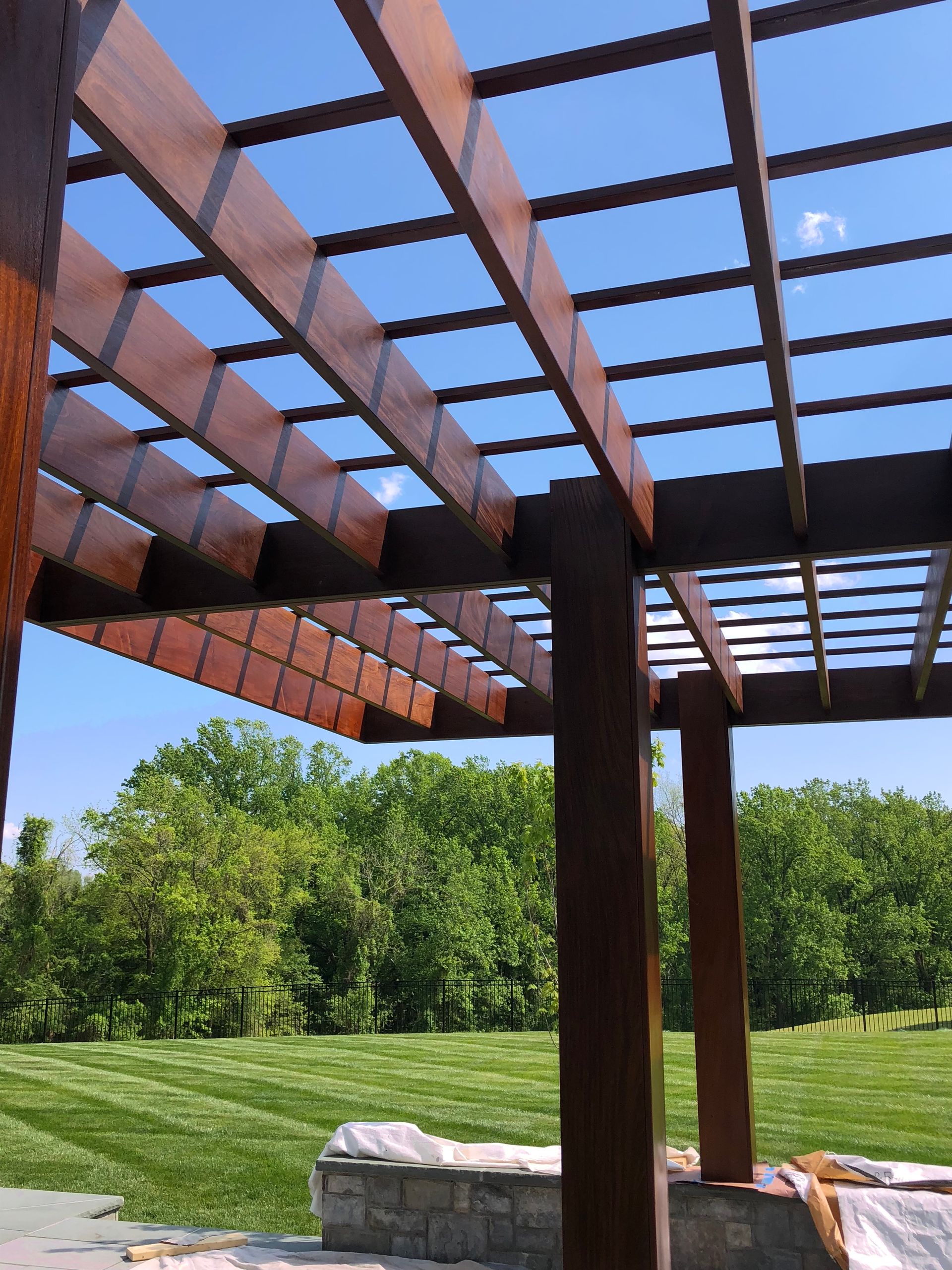 Wooden pergola with latticed roof, over green lawn and trees, blue sky.