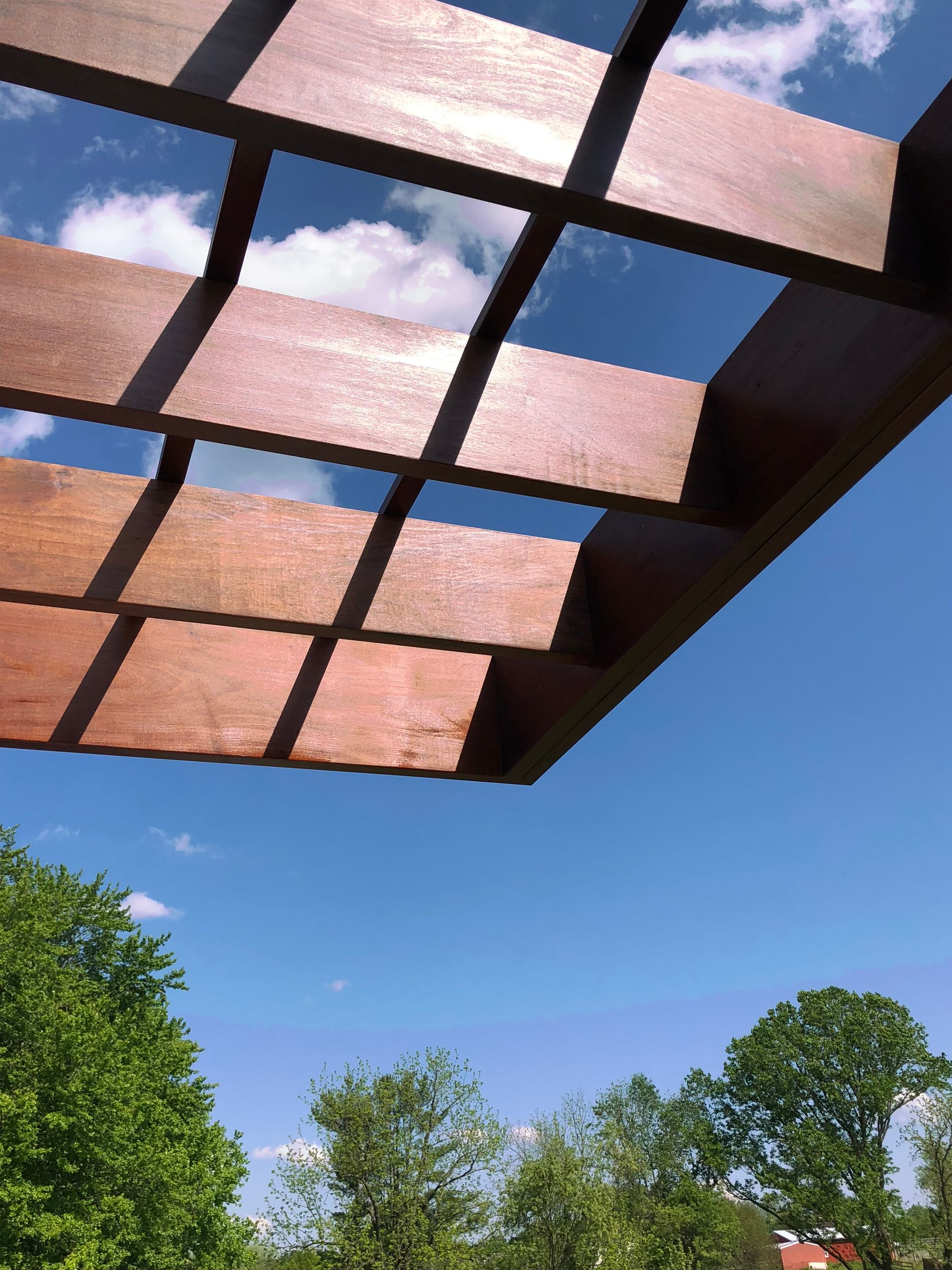 Wooden lattice structure against a blue sky, casting shadows. Trees in the foreground.