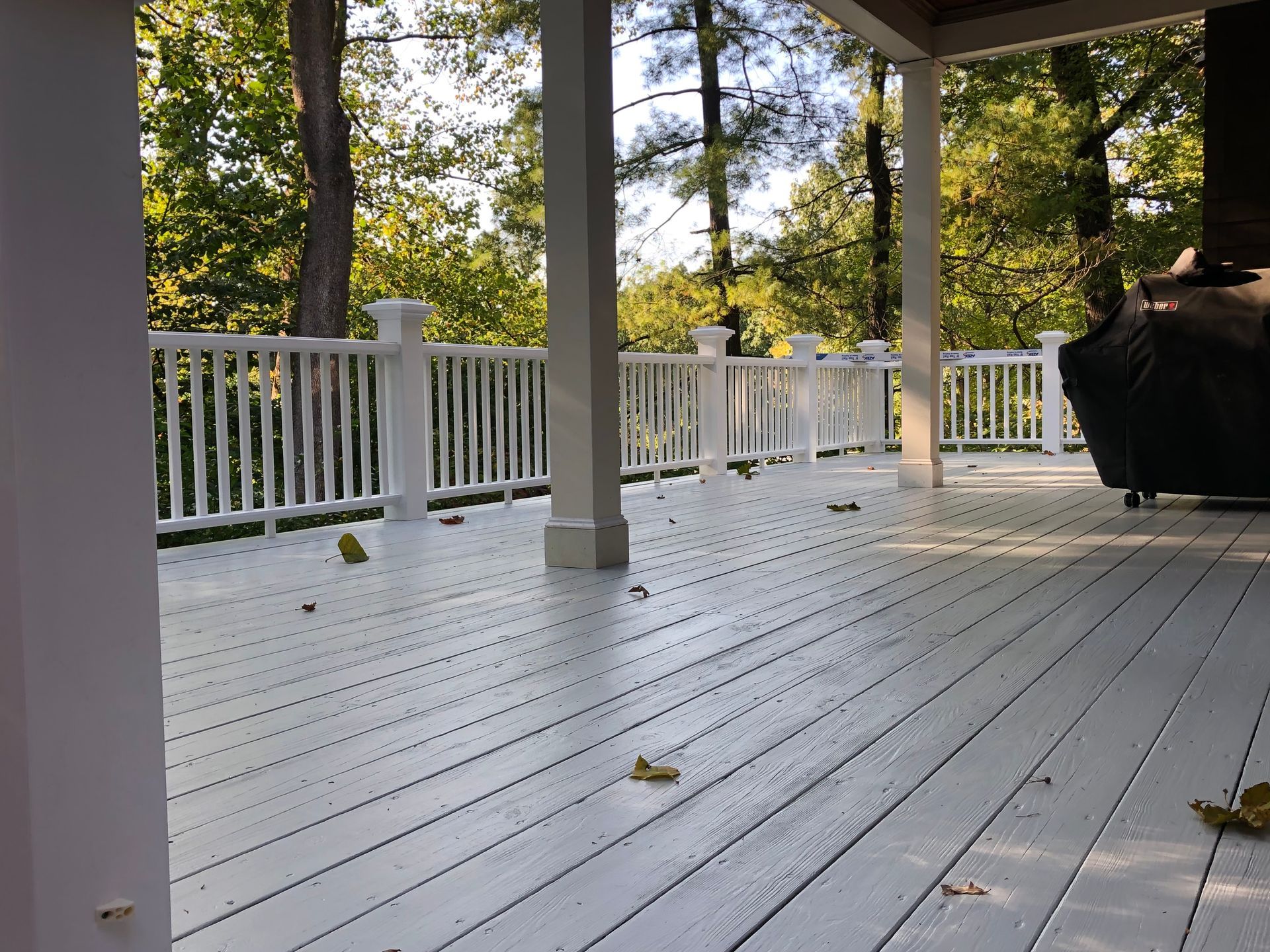 White-painted wooden deck with railing, columns, and a view of trees beyond. Some leaves scattered.