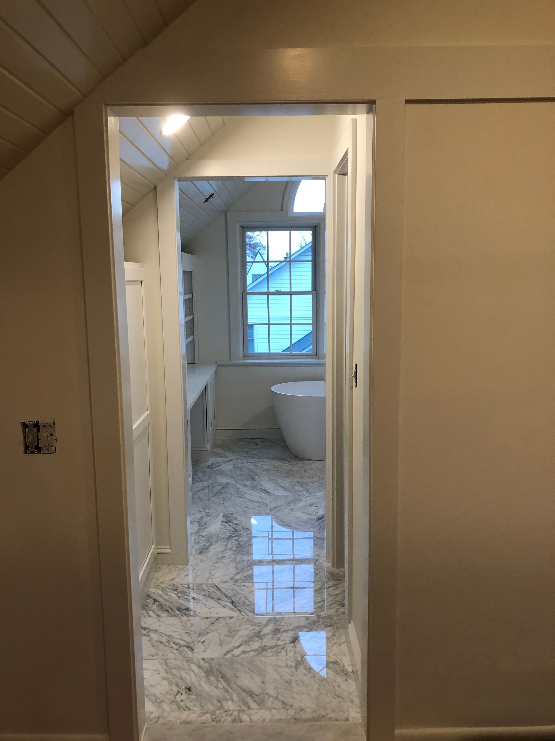 View from a doorway into a white bathroom with marble flooring and a window overlooking a tree-lined street.