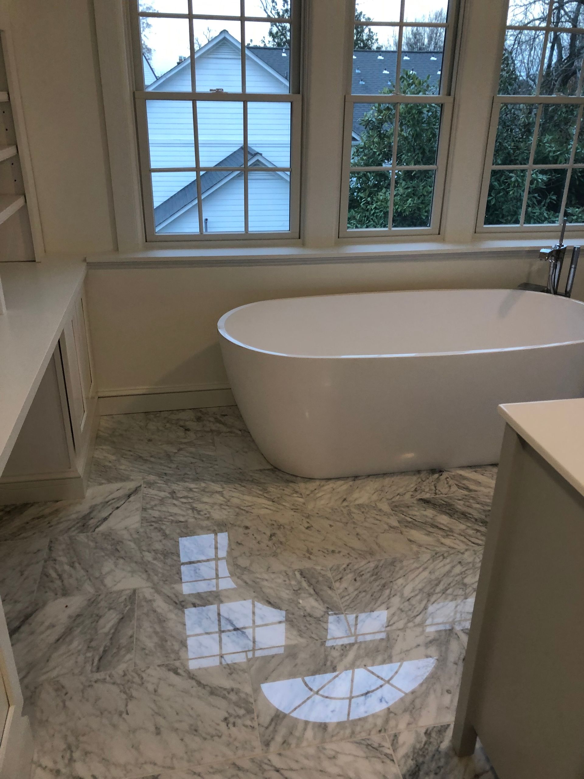 White bathtub in a bright bathroom with marble tile floor and windows overlooking houses.