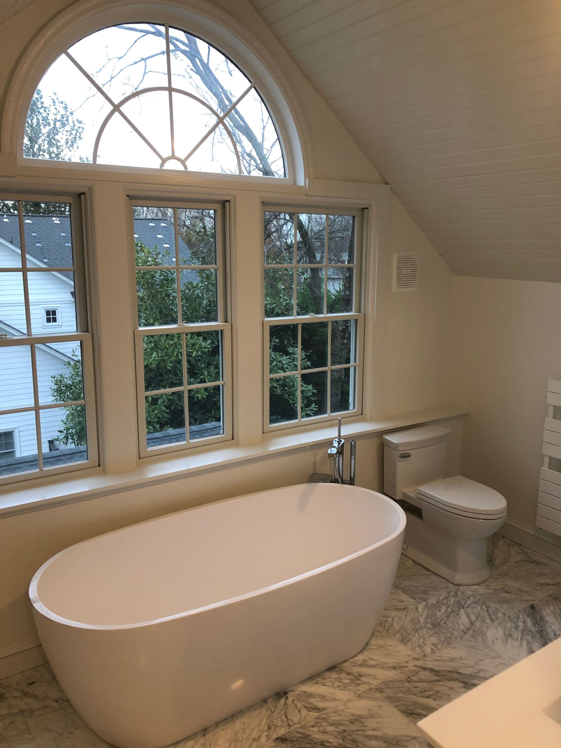 Bathroom with a white oval bathtub, toilet, and windows overlooking greenery. The walls are light.