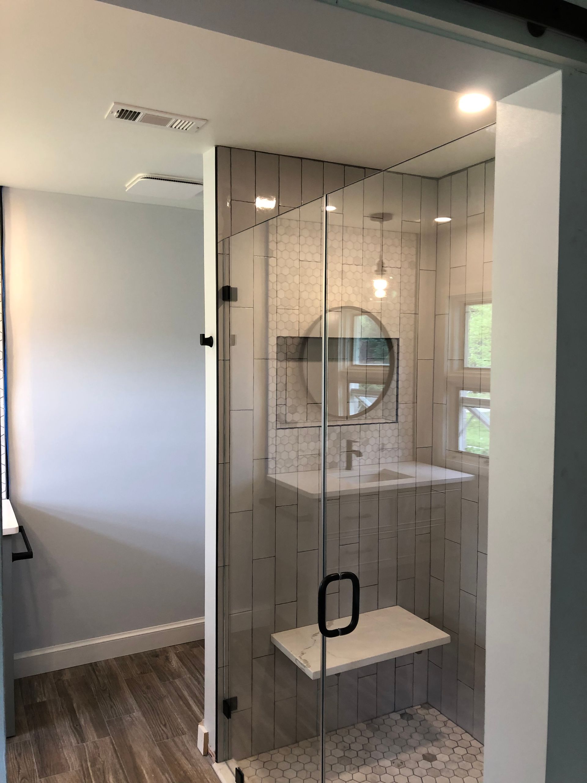 Modern bathroom with glass shower, light gray tile, white sink and bench, recessed lighting, and barn door entry.