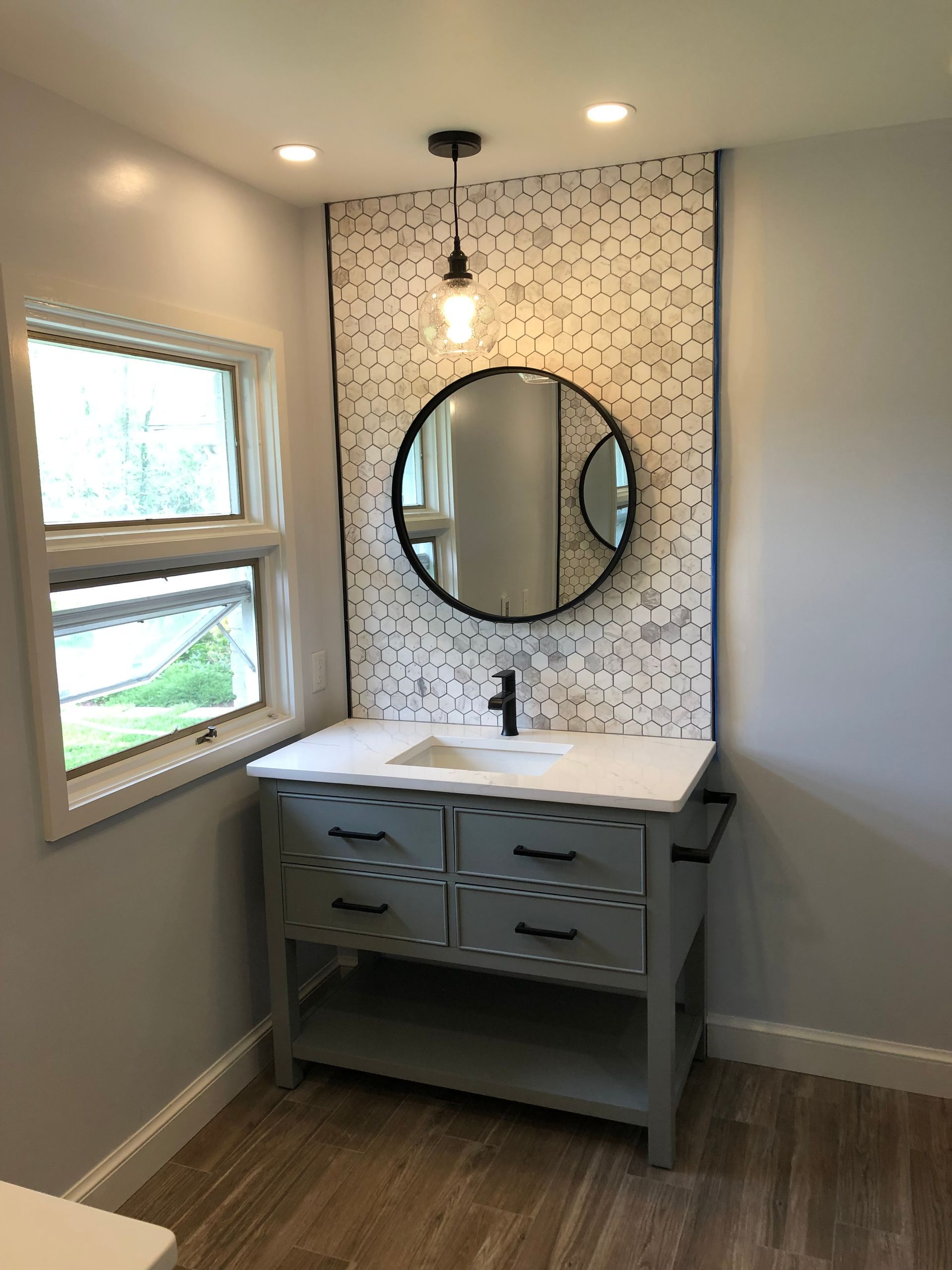 Bathroom with gray vanity, round mirror, and patterned backsplash. Window to the left.