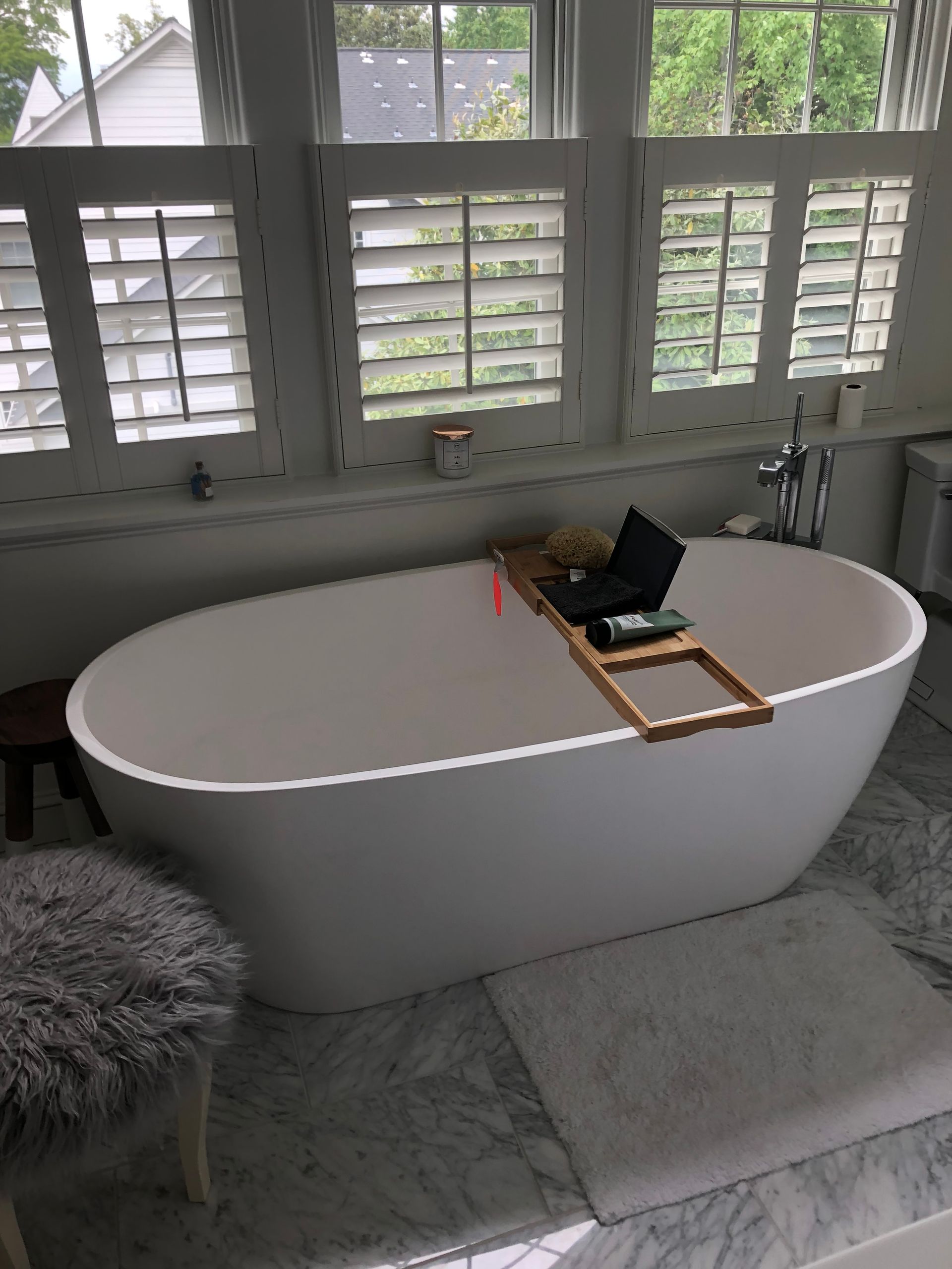 White oval bathtub with a wooden tray holding a laptop, in a bathroom with window shutters.