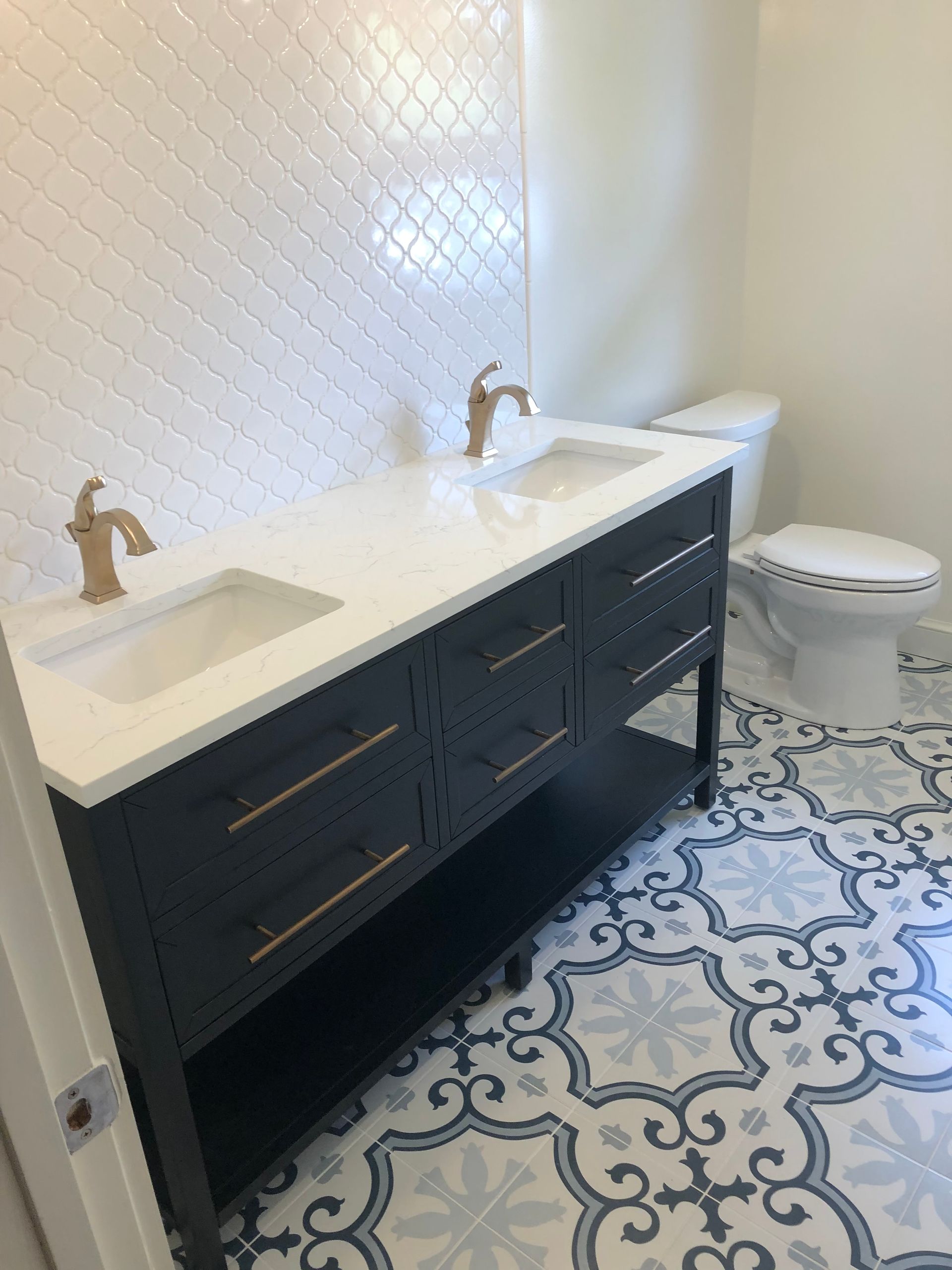 Bathroom with dark blue double vanity, white countertop, gold fixtures, patterned floor, and white tiled wall.