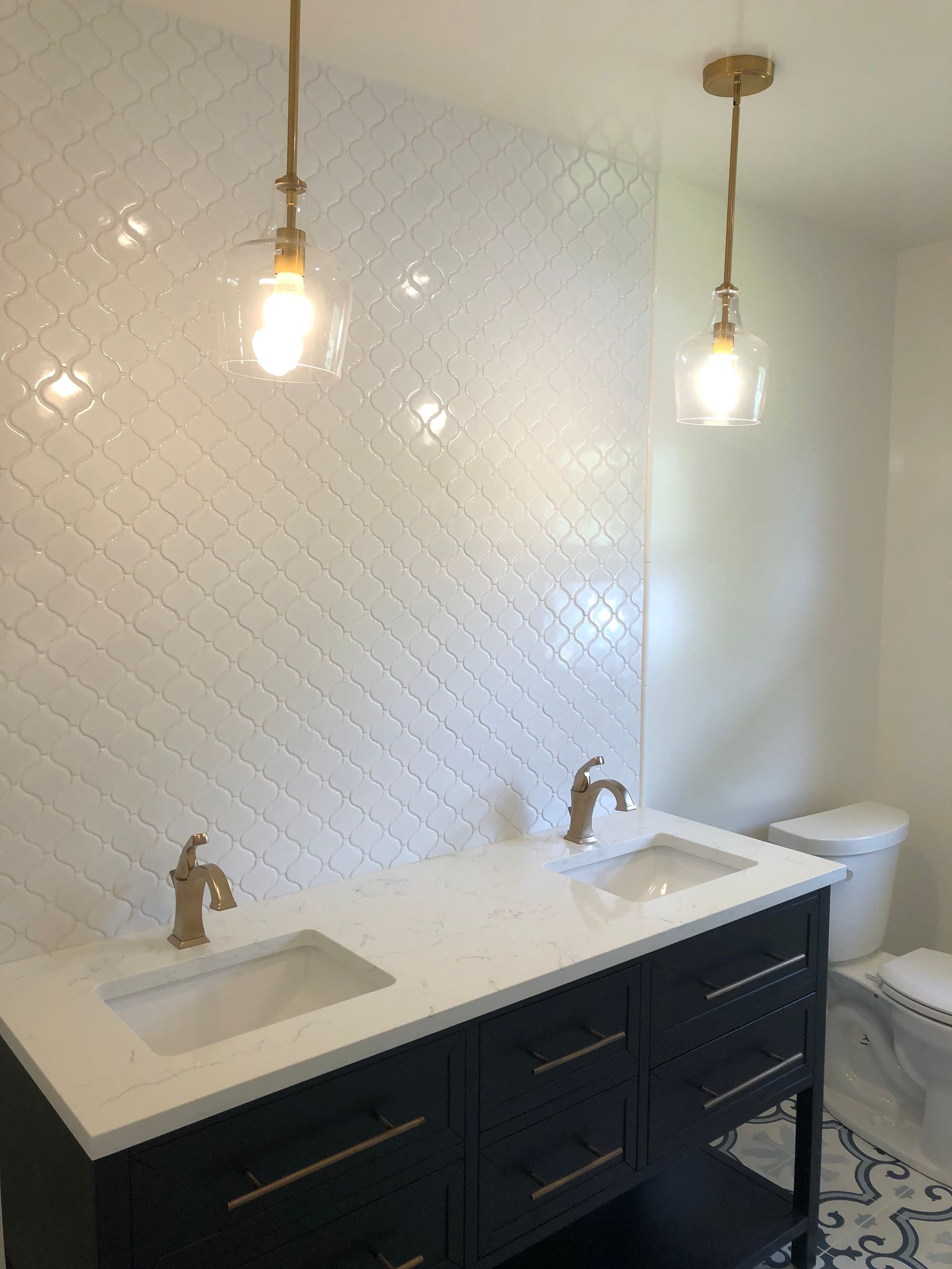 Bathroom with black vanity, white countertop, two sinks, gold fixtures, white tiled wall, two pendant lights.