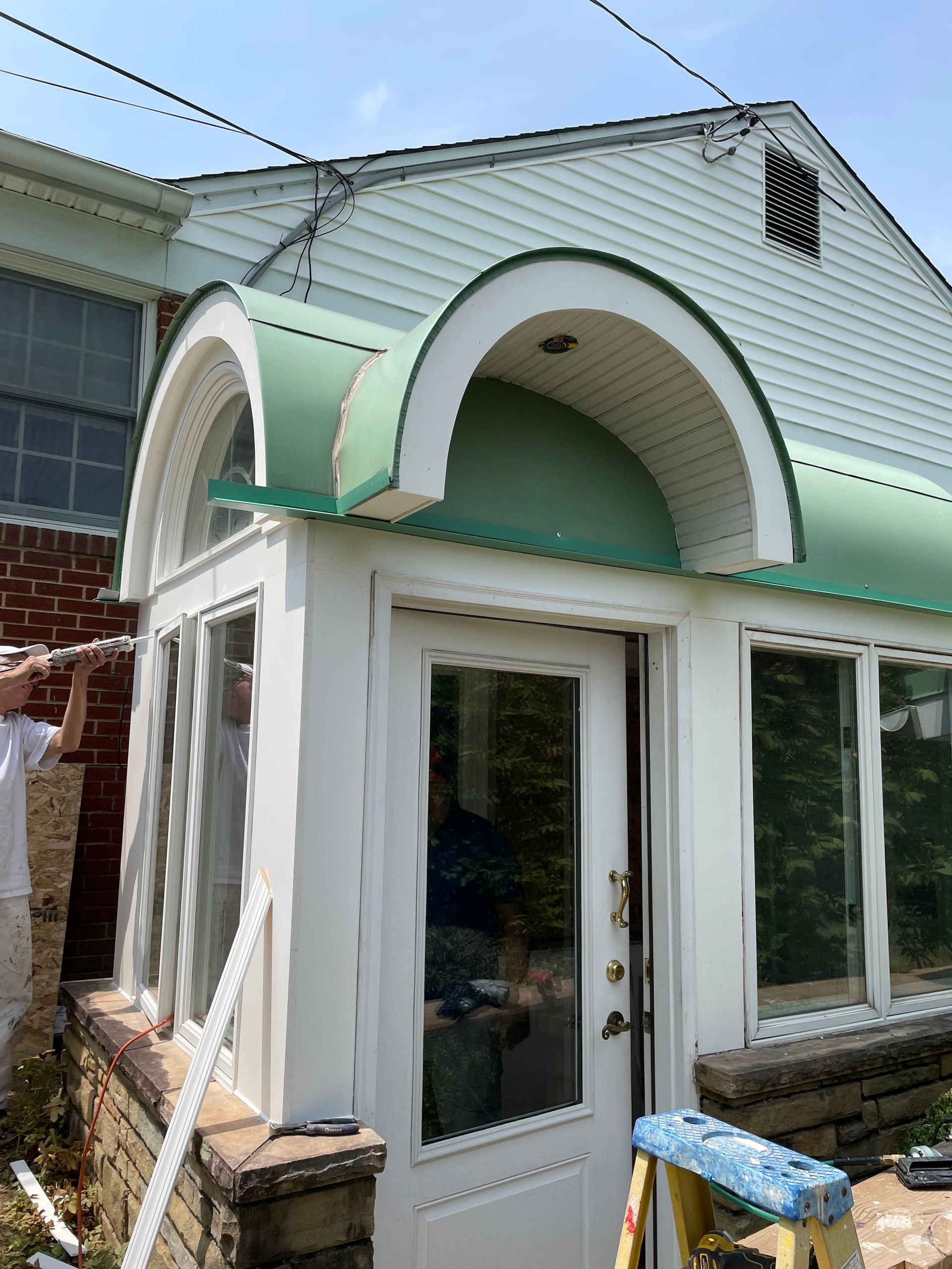 Green-roofed entryway with arched design and glass windows, attached to a house with white siding and a brick base.