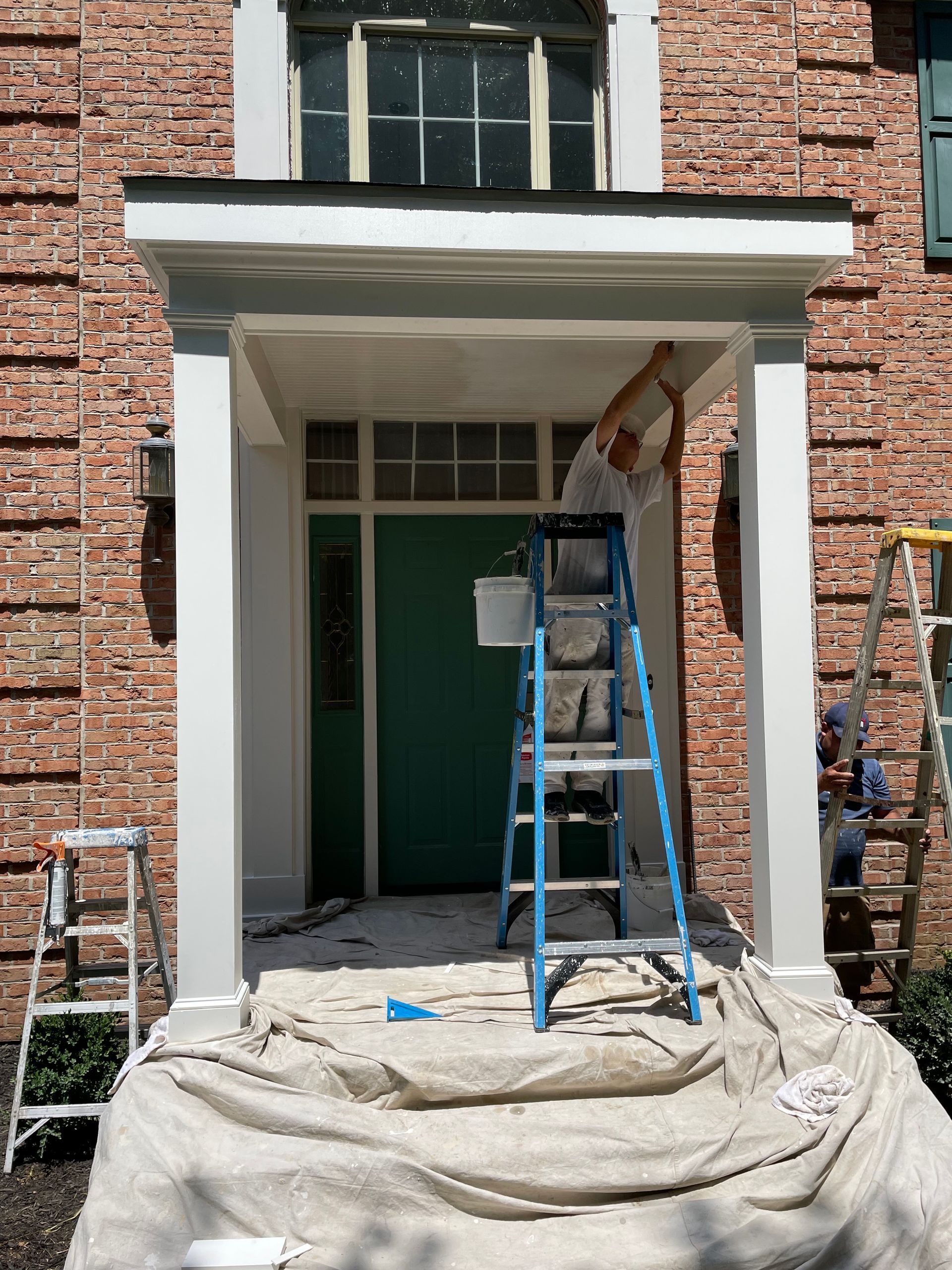 Person on ladder painting a white porch trim. Green door, brick building.