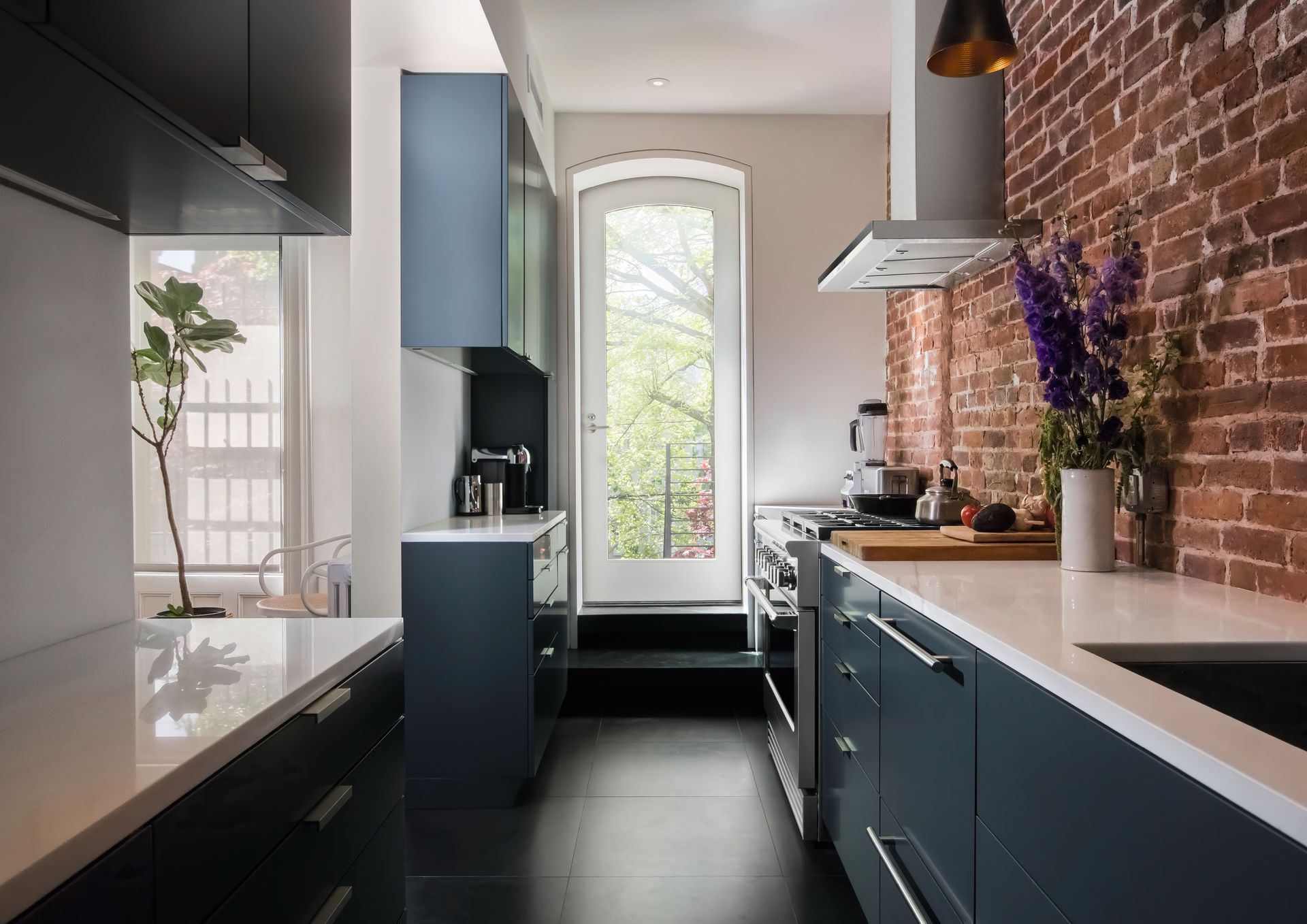 Narrow kitchen with dark blue cabinets, white countertops, brick wall, and a door leading outside.