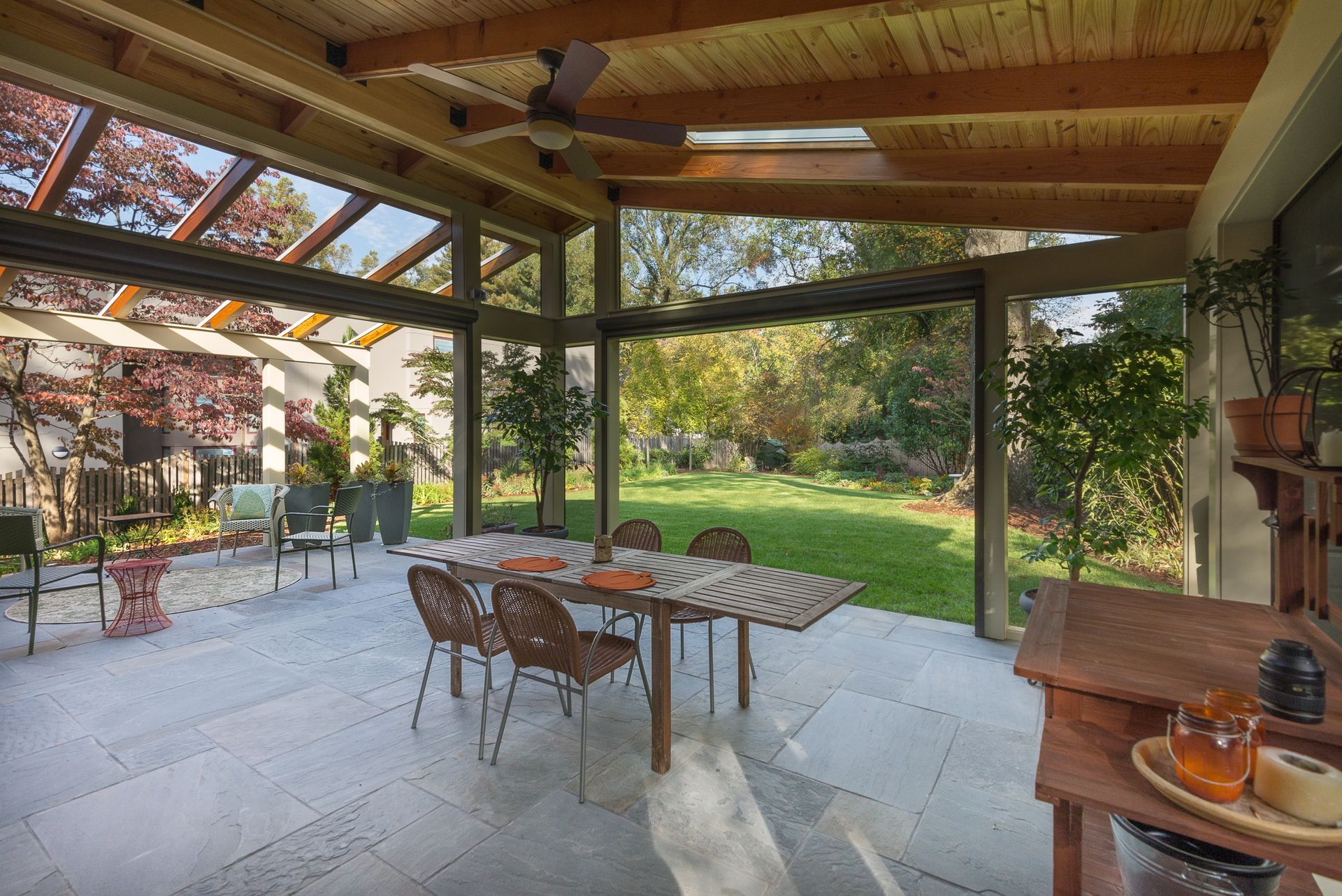 Covered patio with dining table overlooking a green yard and trees; wooden beams, blue-gray tile floor.