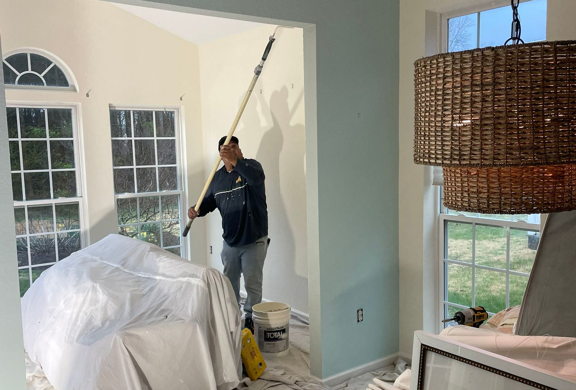 Person paints a room with a roller, standing near windows. Furniture is covered with plastic sheeting.