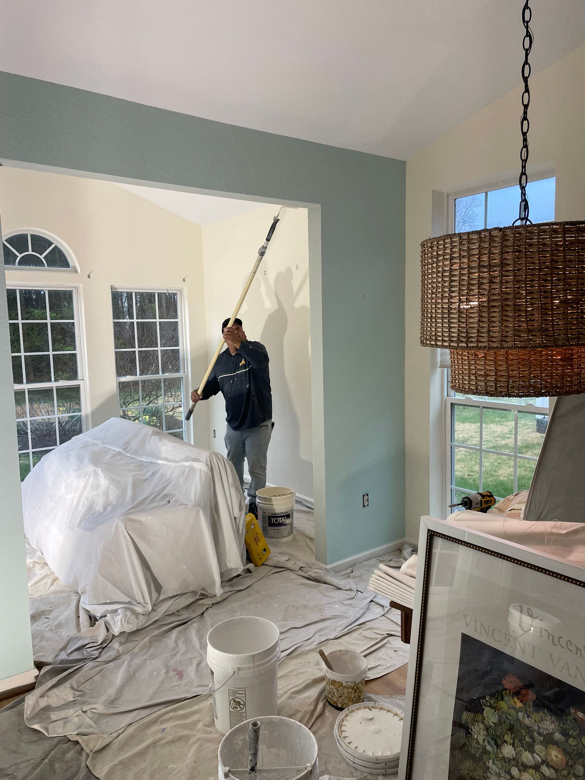 Man painting a room with blue-green accent wall and beige walls. Room has multiple windows and a large pendant light.