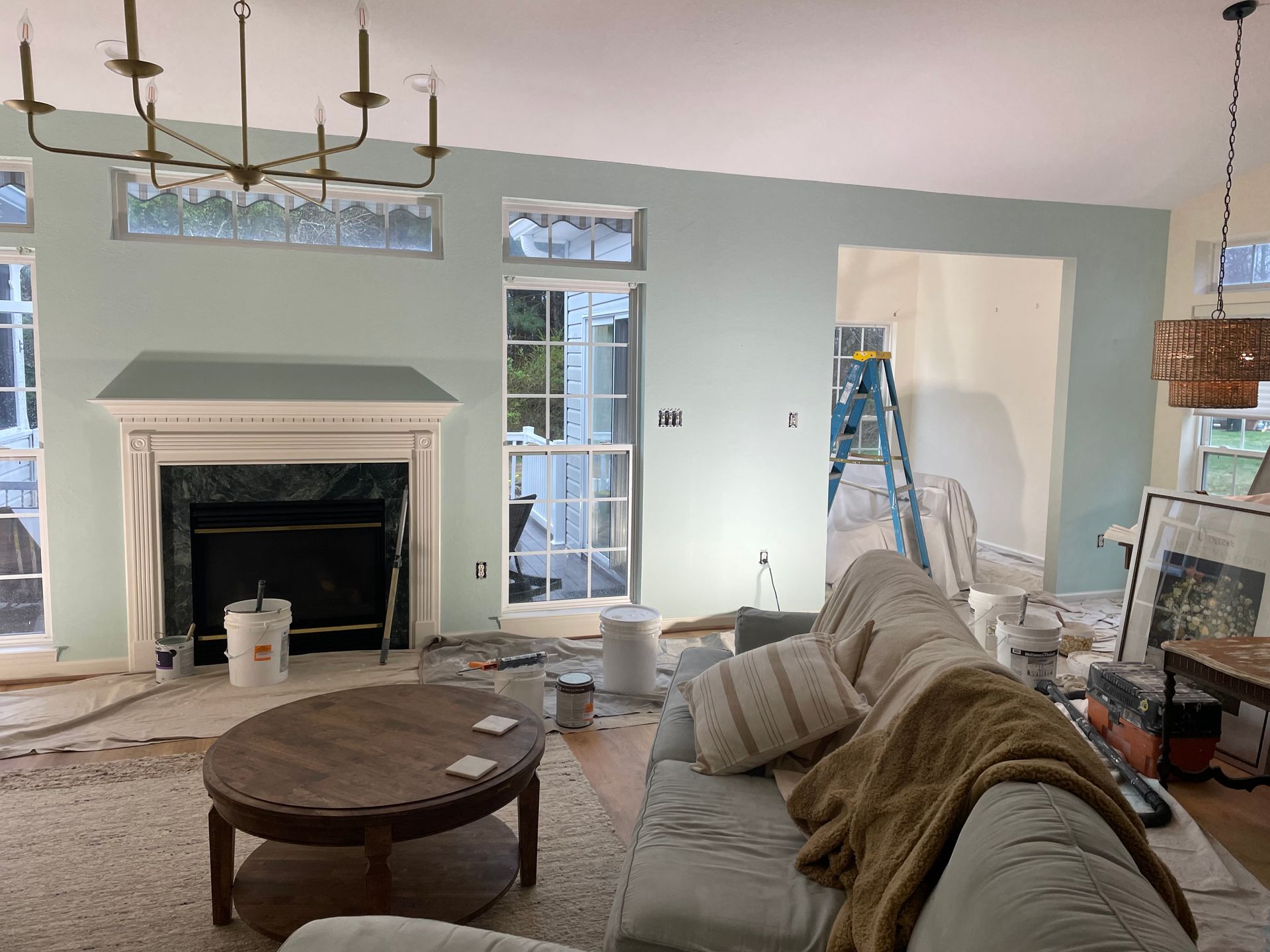 Living room with blue-green walls during renovation. Fireplace, windows, sofa, and painting supplies visible.