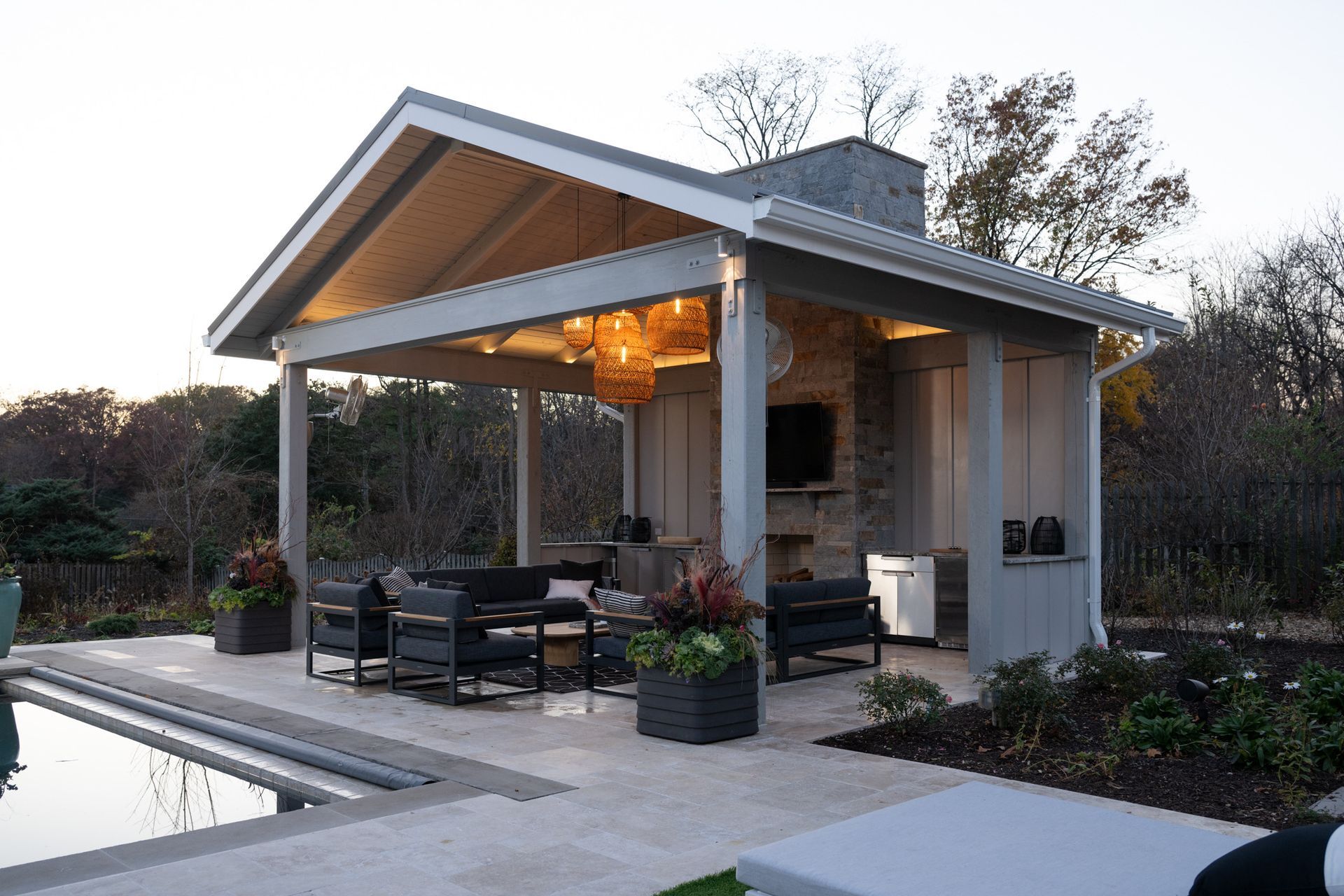 Poolside outdoor living space with gray-painted wood pavilion, fireplace, seating, and a decorative light fixture.