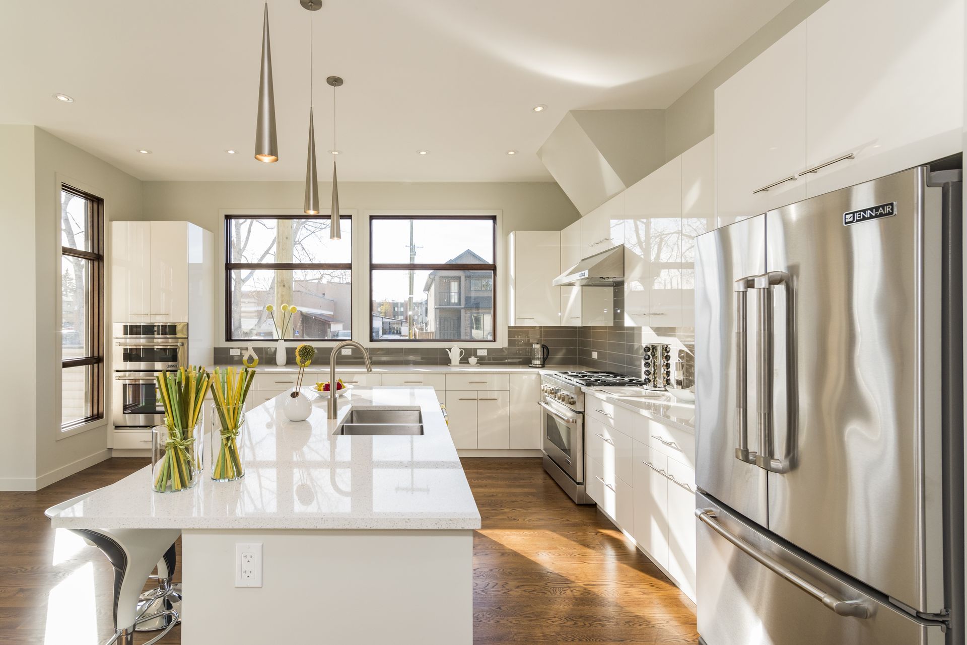 Modern white kitchen with island, stainless steel appliances, and large windows.