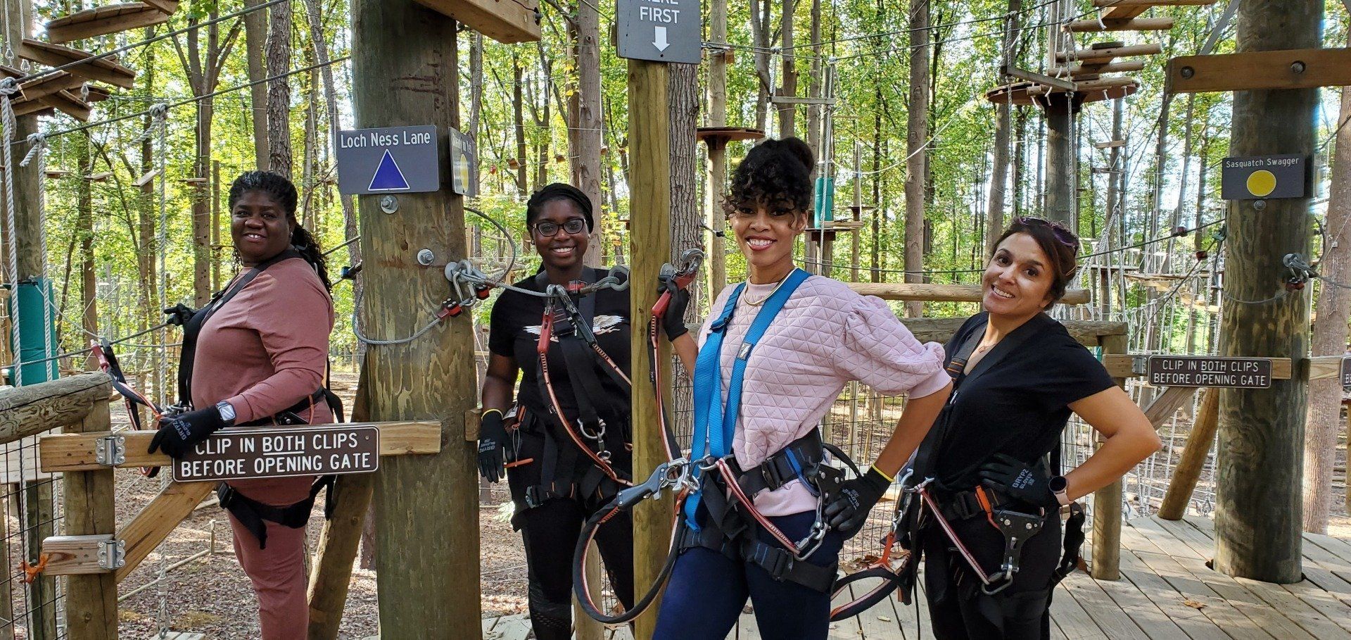 Four people wearing harnesses stand on a wooden platform in a forest adventure course.