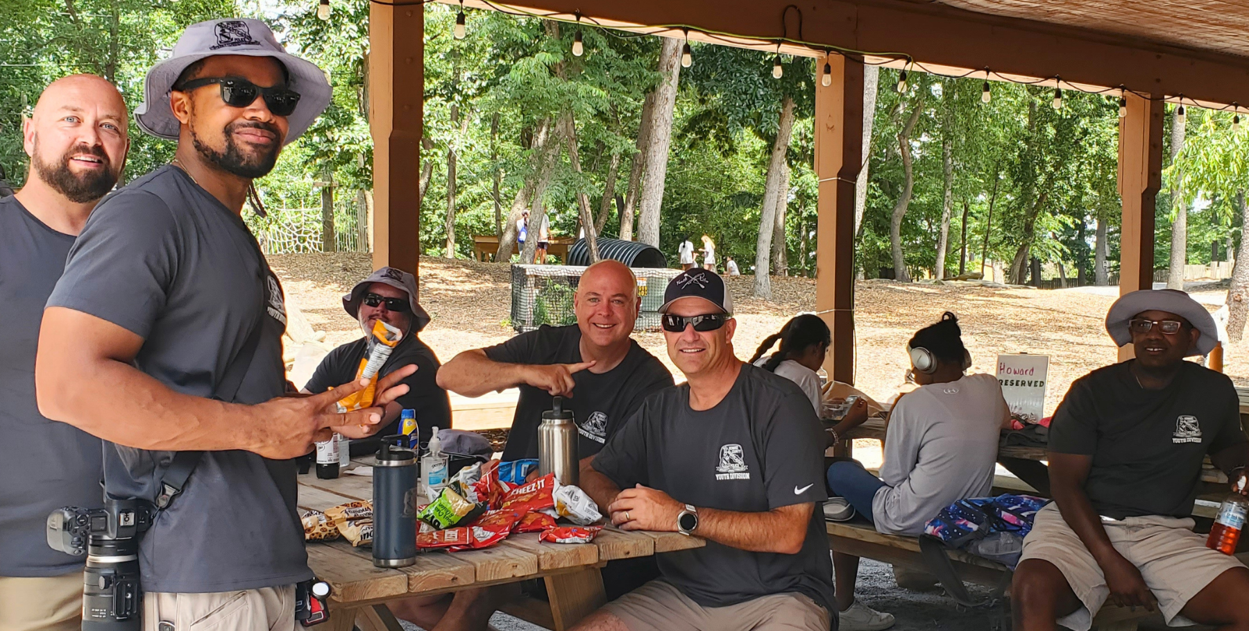 Group of people socializing under a park pavilion, seated at picnic tables with drinks and snacks.