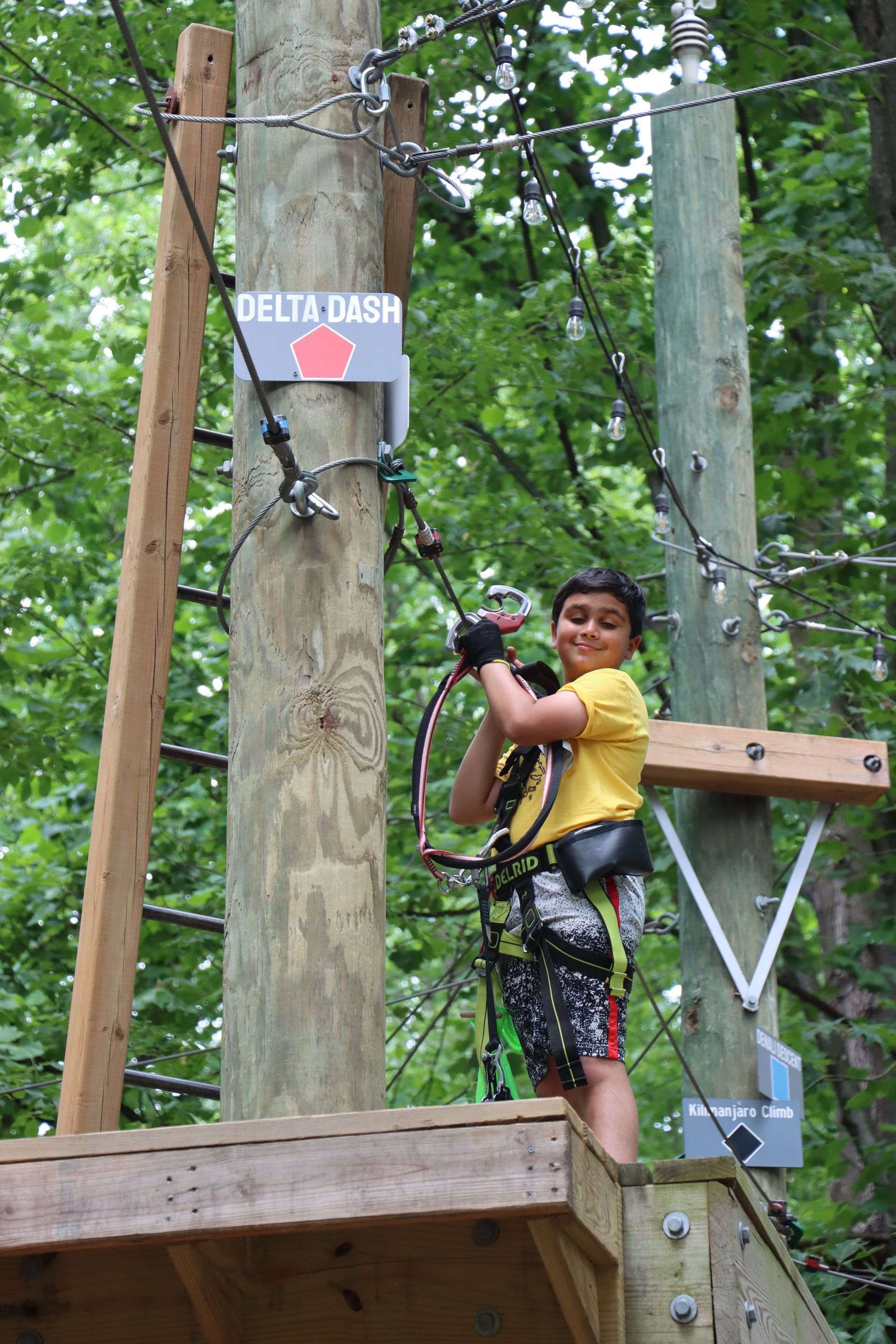 A young boy is standing on top of a wooden platform on a ropes course.