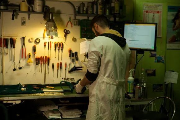Persona al lavoro su un banco da lavoro ingombro di attrezzi in un'officina, con utensili appesi al muro e un monitor nelle vicinanze.