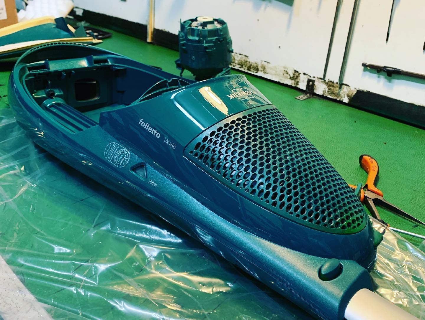 Scafo di kayak blu in un'officina, con ponte in rete nera e attrezzo arancione nelle vicinanze su un pavimento verde.