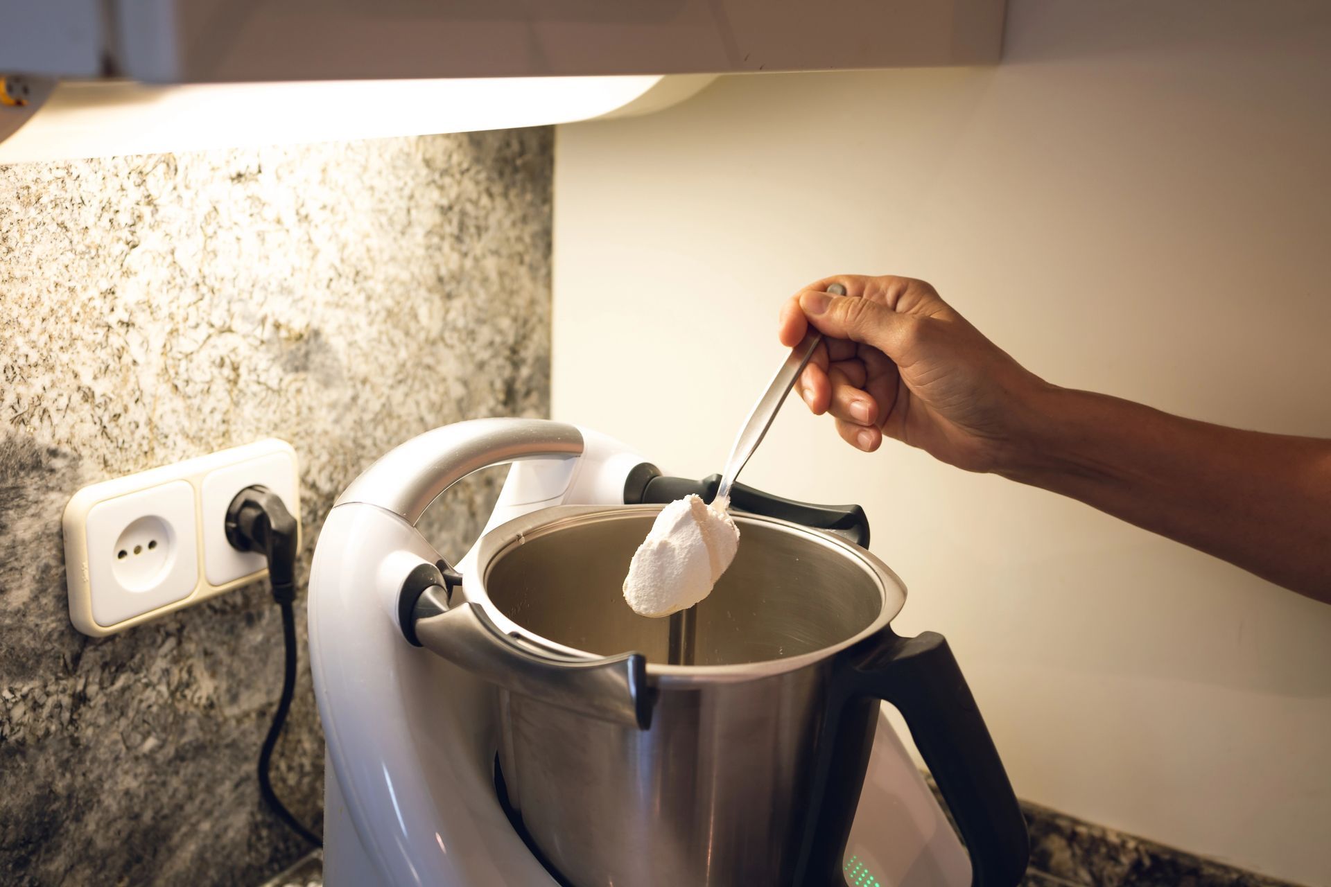 Una mano che tiene un cucchiaio sopra la ciotola di un'impastatrice da cucina vicino a una presa a muro.