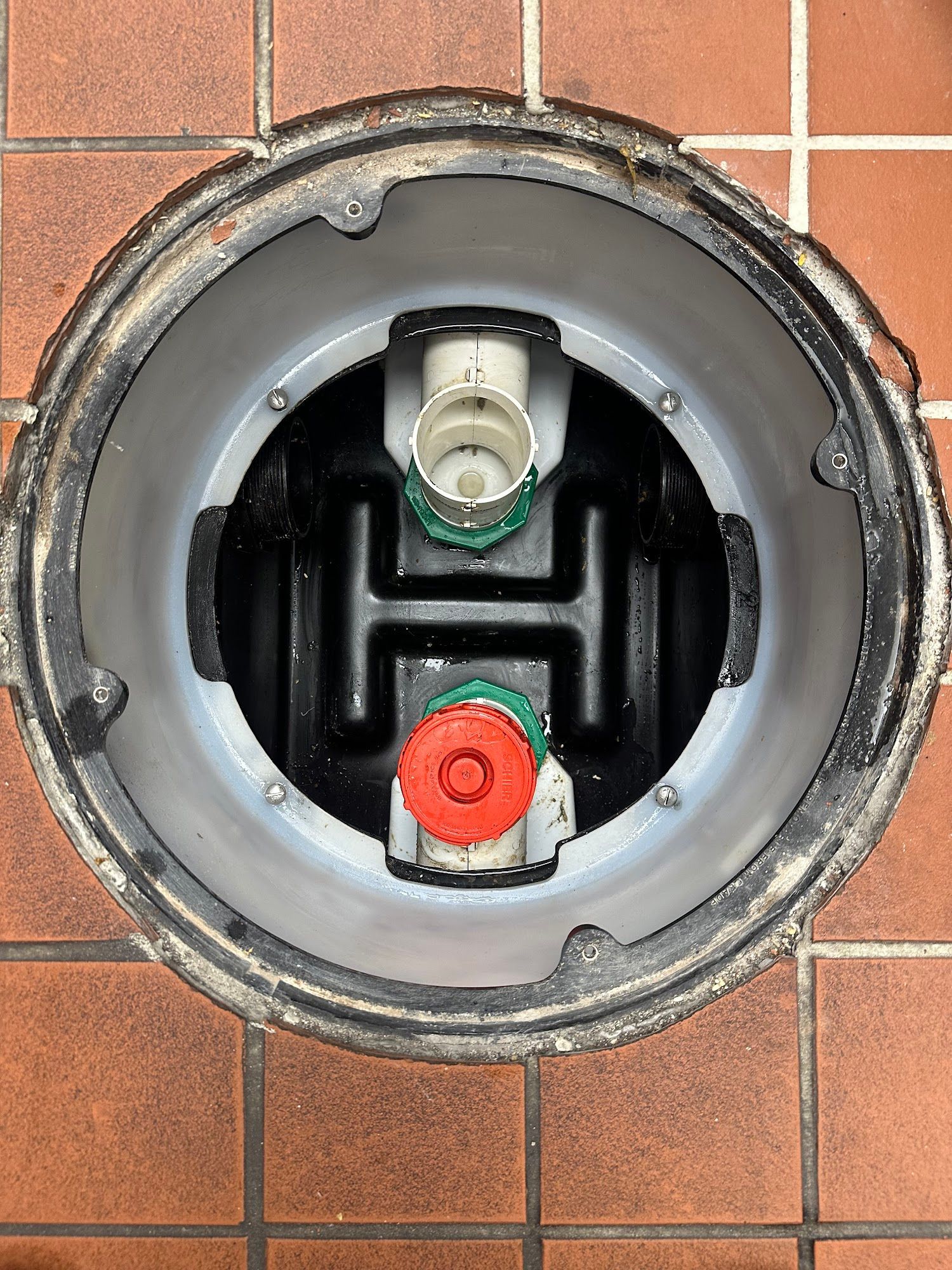 Open manhole with black plastic structure inside, surrounded by a metal rim on brown brick.