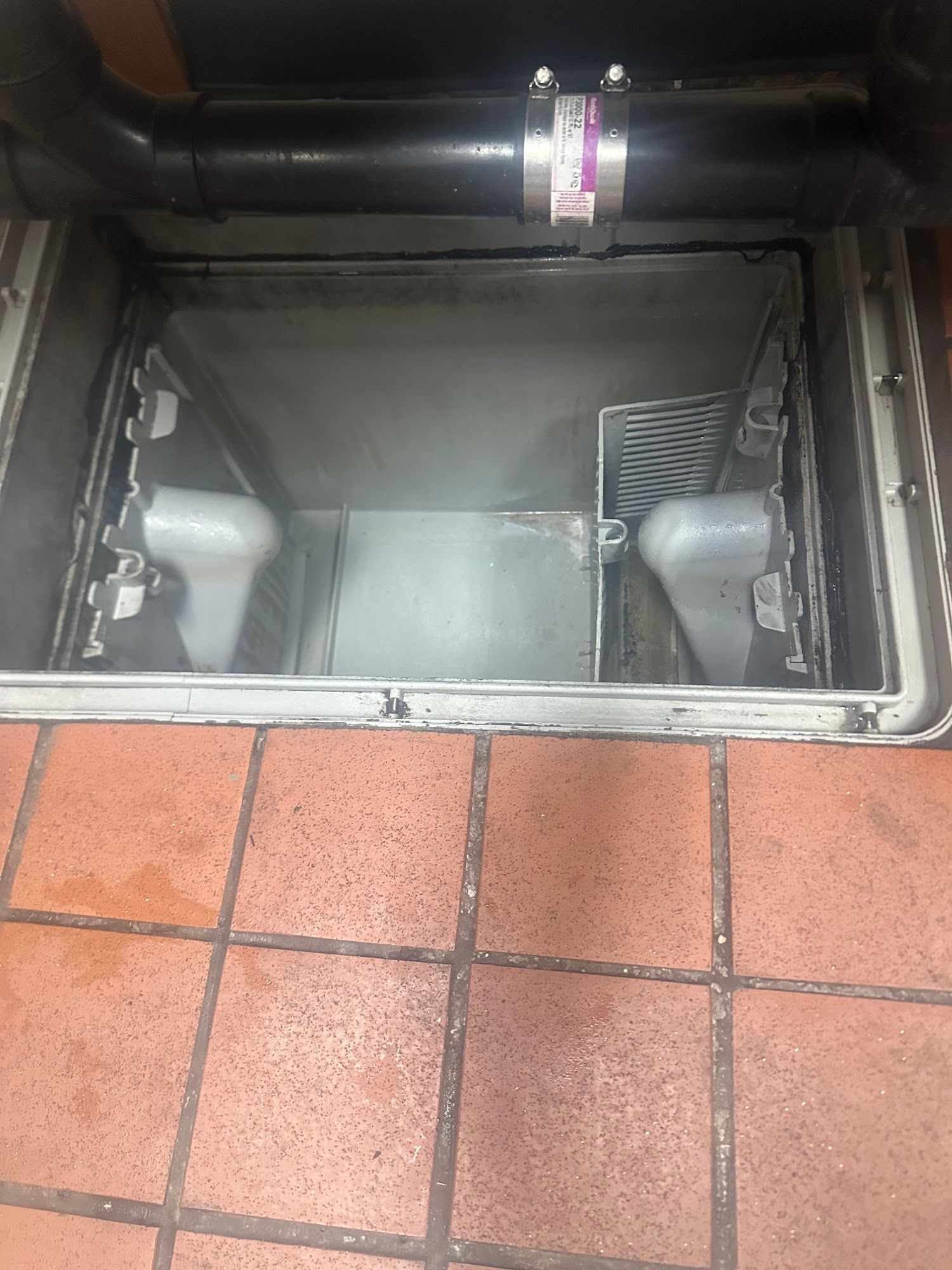 Open, rectangular floor drain with metal components. A black pipe is above the drain, and the floor is red tile.