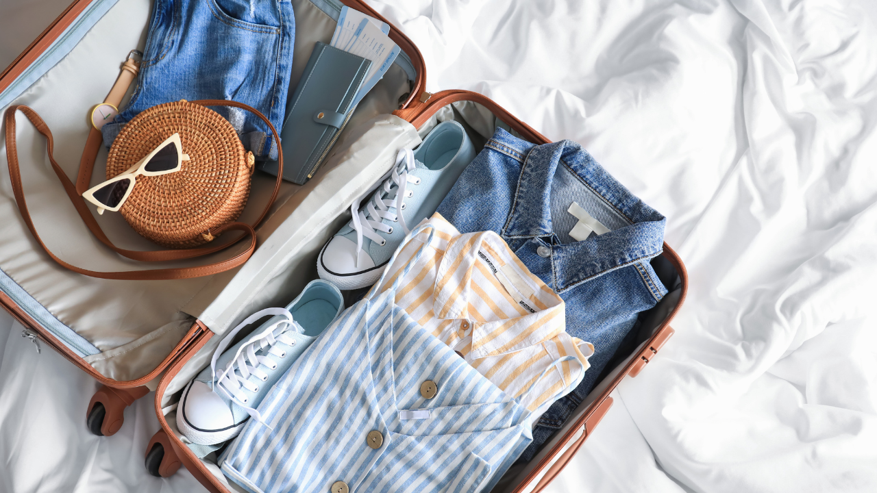 Open suitcase with folded clothes, jeans, sneakers, and a brown handbag on a white bed.