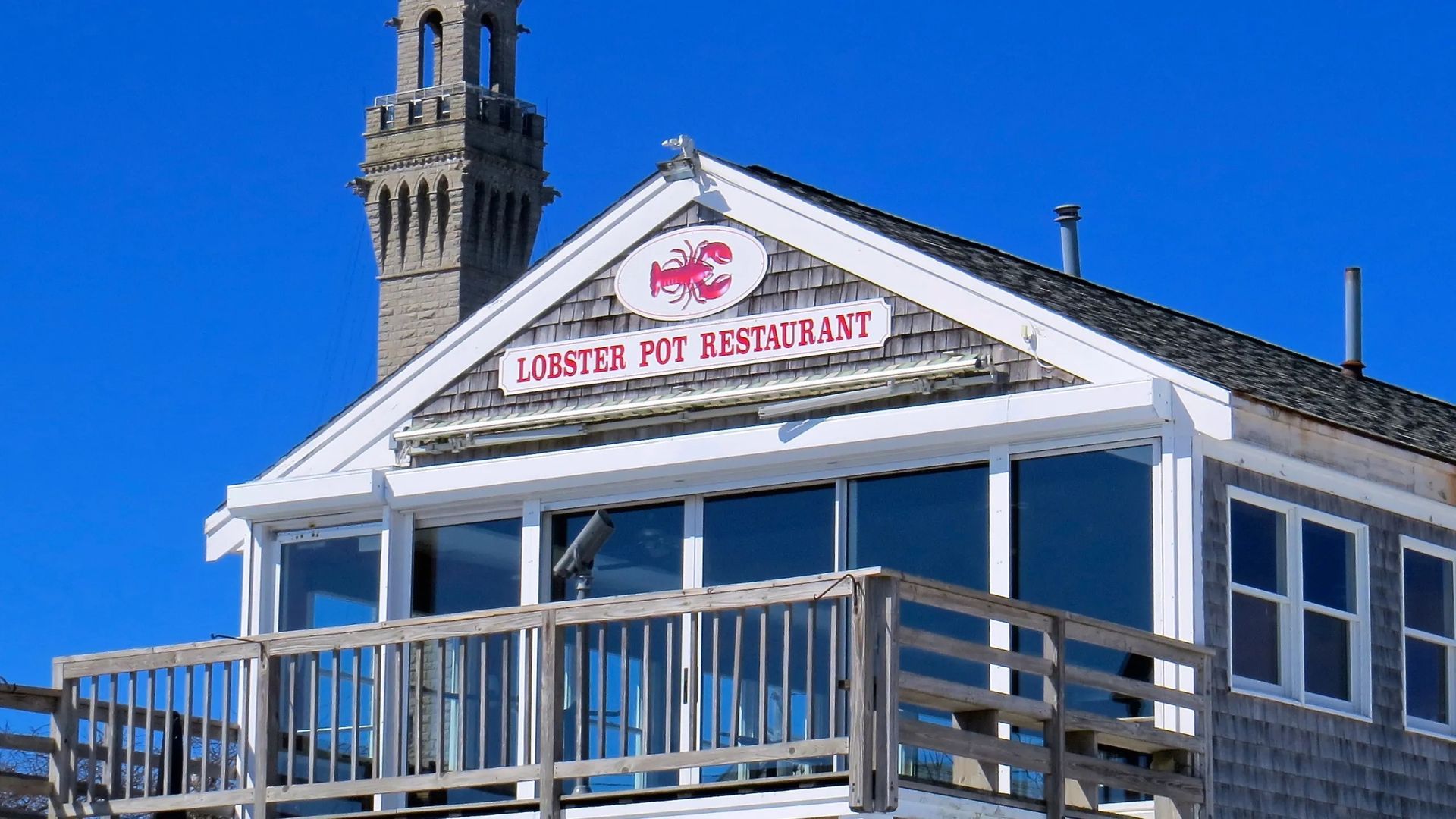 A weathered, gray-shingled building with a lobster sign and a wooden deck sits against a clear blue sky.