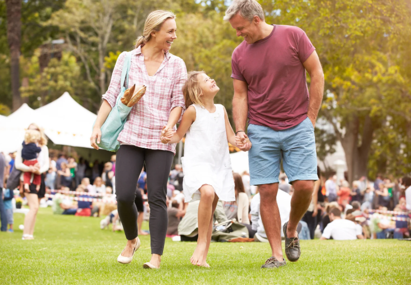 A family walks hand-in-hand across a grassy park with event tents and a crowd in the background.