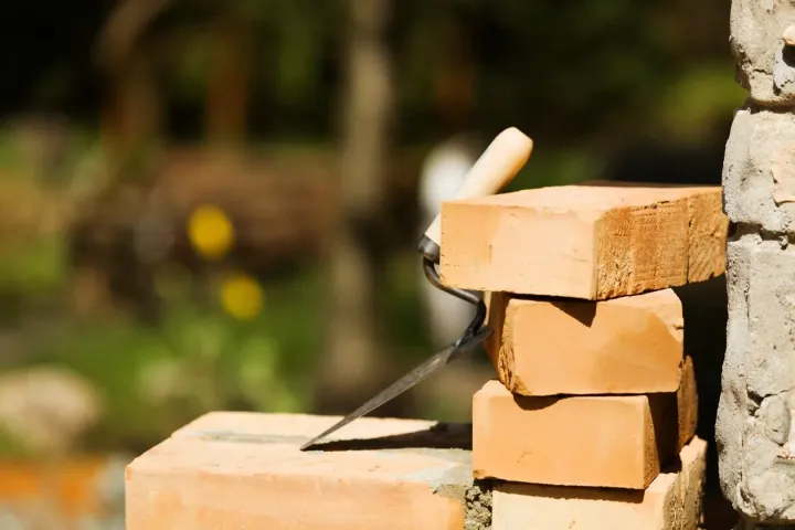 A metal trowel rests between a stack of red clay bricks outdoors in a garden setting.