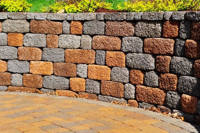 A curved stone retaining wall featuring rectangular blocks in shades of tan and gray, set against a paved patio.