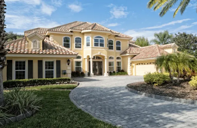 A two-story, cream-colored stucco home with a brown tile roof and a stone driveway on a sunny day.