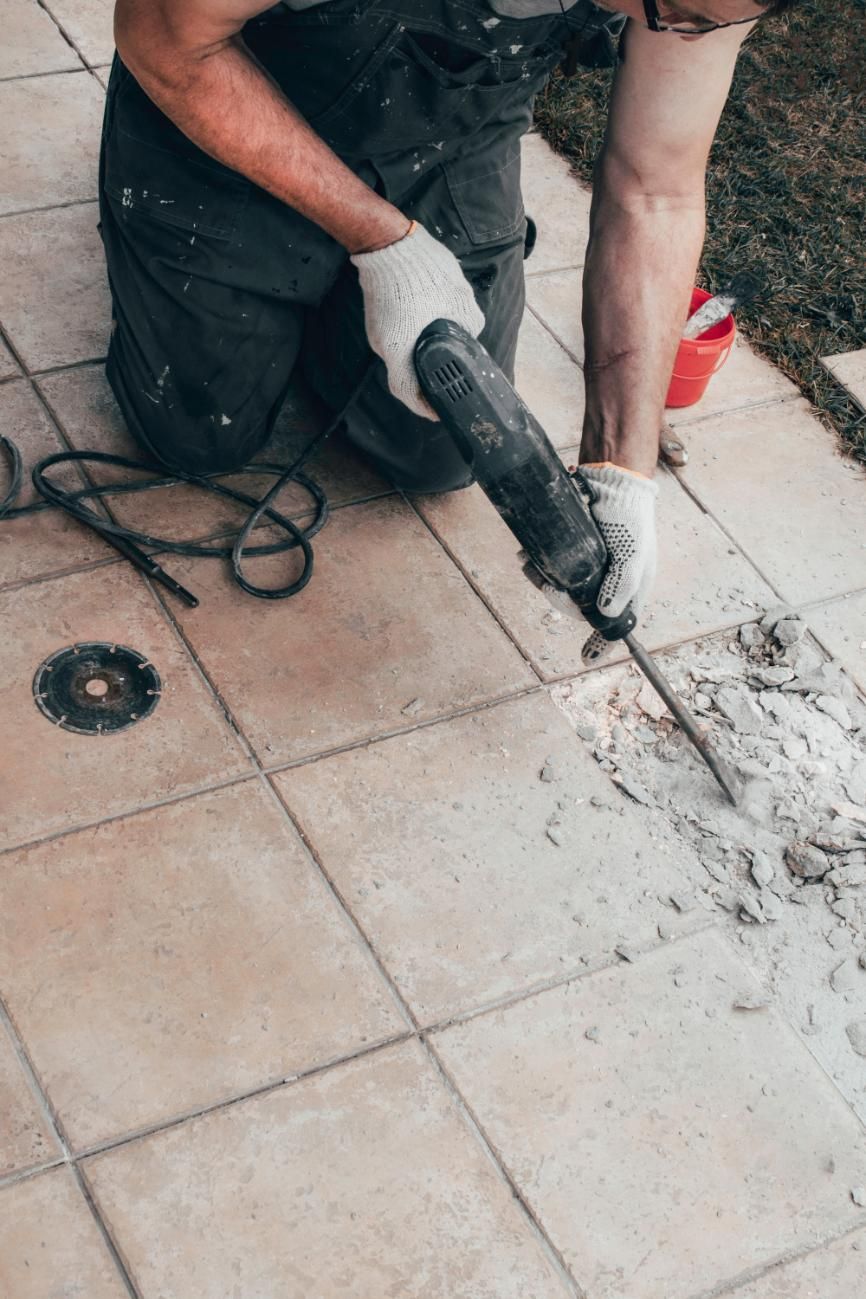 A Man Is Using a Hammer to Drill a Hole in A Tile Floor — TNT Floor Removal & Demolition in Tewantin, QLD