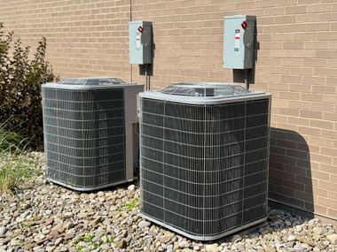 Two outdoor HVAC units beside a brick wall with electrical disconnect boxes above them