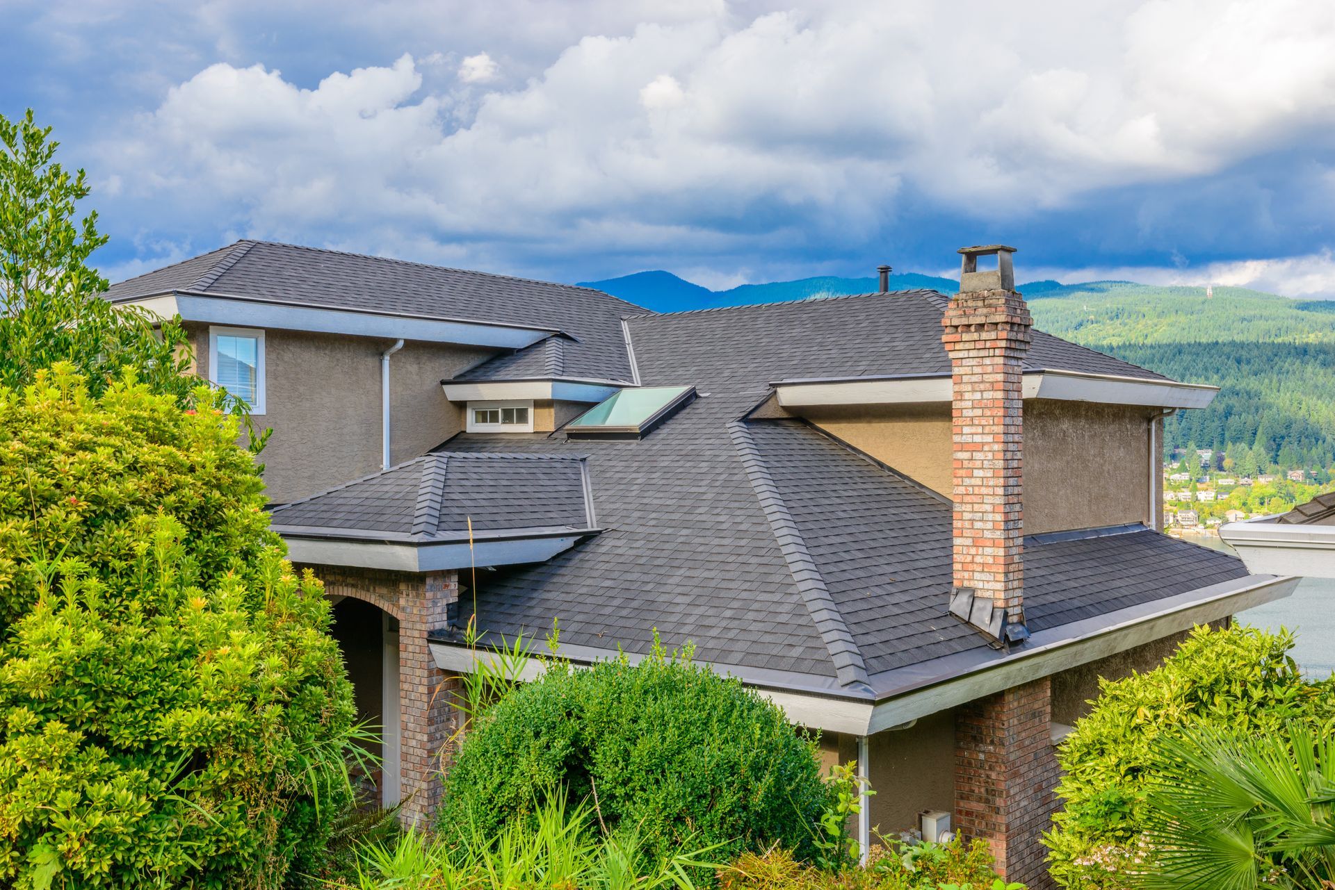 A modern, two-story house with a grey shingled roof, brick chimney, and skylight, set against mountains and blue sky.