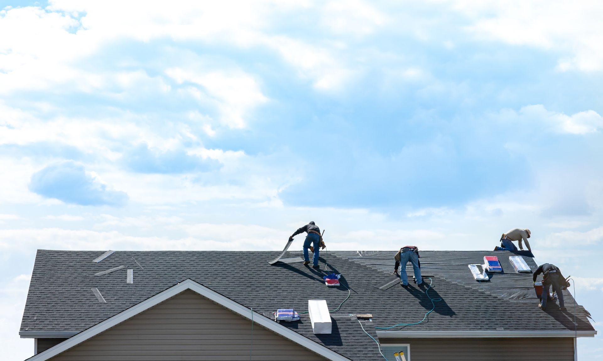 Four workers wearing safety gear install dark asphalt shingles on a suburban house roof under a cloudy sky.