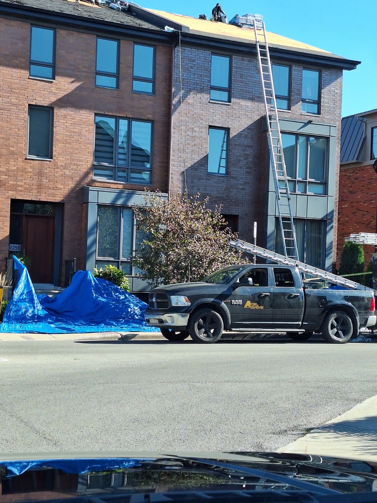 Une camionnette sombre garée devant des maisons de ville en briques, avec des ouvriers sur le toit utilisant une échelle pour des travaux de construction.