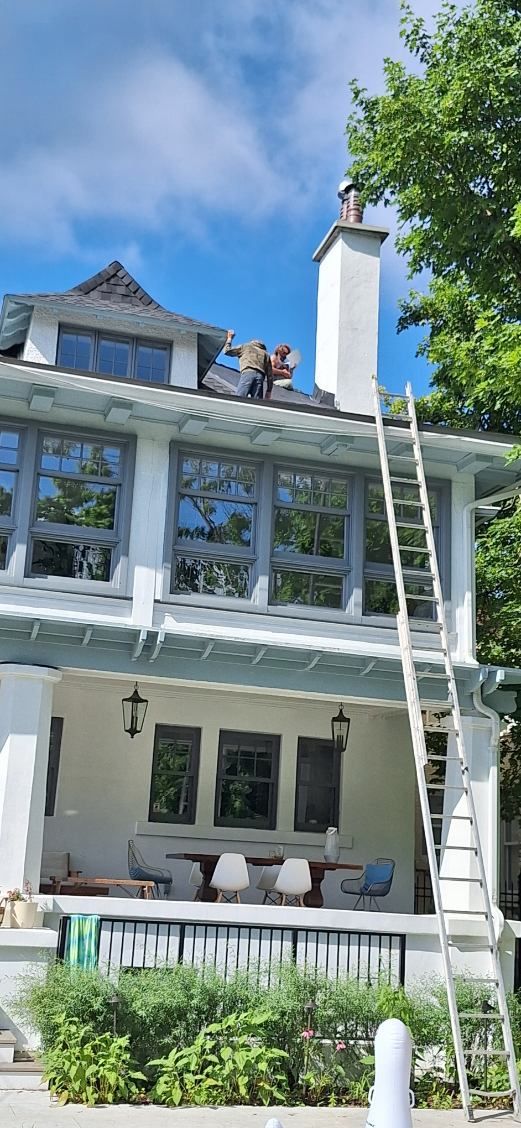 Une grande échelle est appuyée contre la façade d'une maison blanche, menant à une personne effectuant des travaux d'entretien sur le toit.