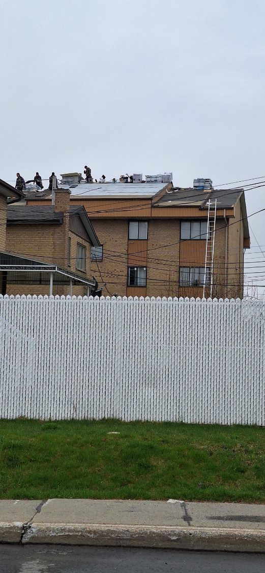 Construction workers repairing the roof of a multi-story brick apartment building, viewed over a white privacy fence.
