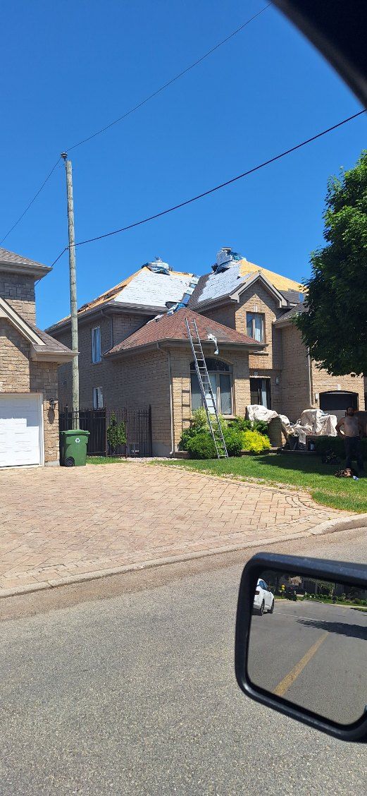A house under construction with exposed roofing sections and a ladder, viewed from a car mirror on a sunny day.